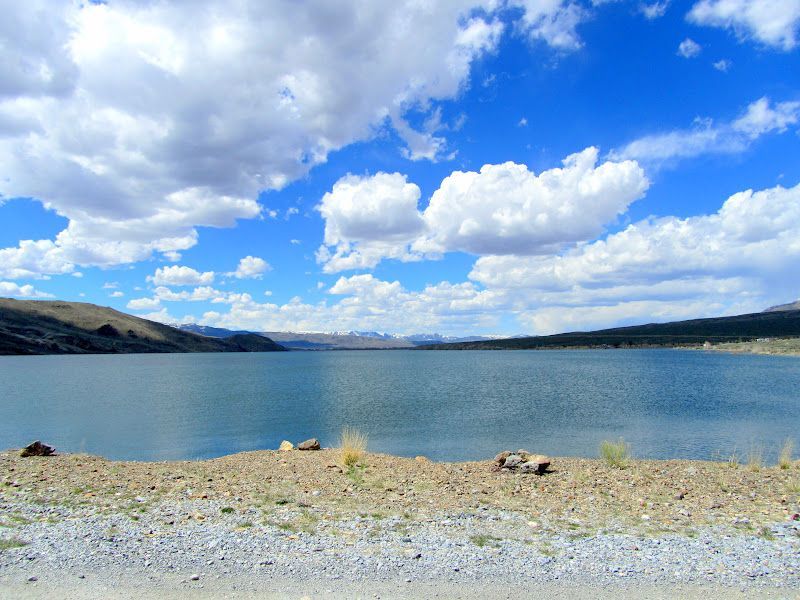 A clear blue lake under a vast, cloud-filled sky, bordered by rocky, dry shoreline and distant mountains.