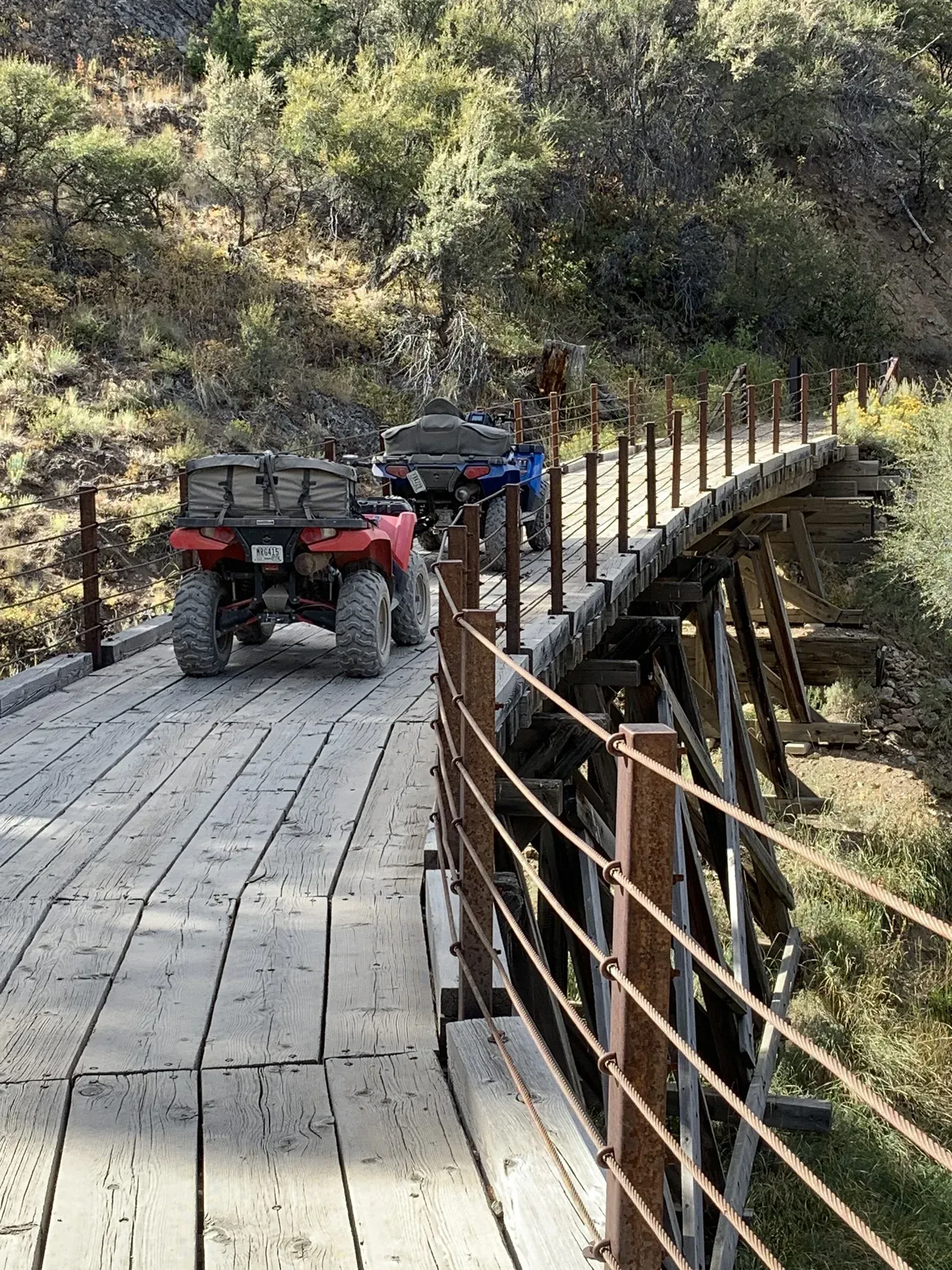 Two ATVs travel across a weathered wooden bridge with metal railings through a wooded area.
