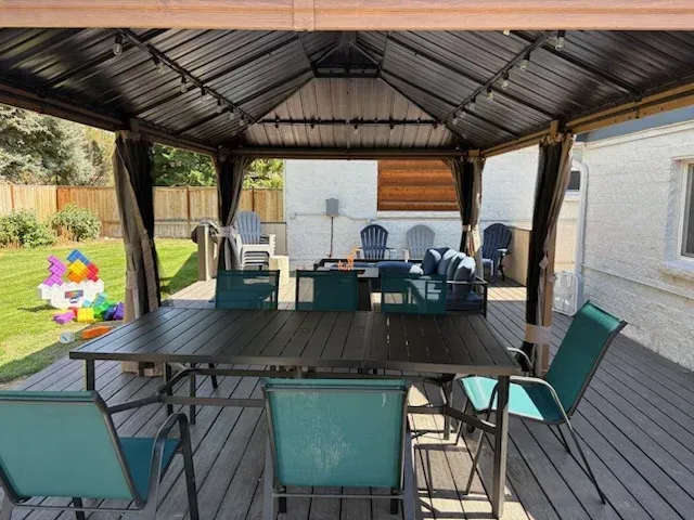 A patio deck featuring a dark metal gazebo over a rectangular table with teal chairs, with a grassy lawn in the background.