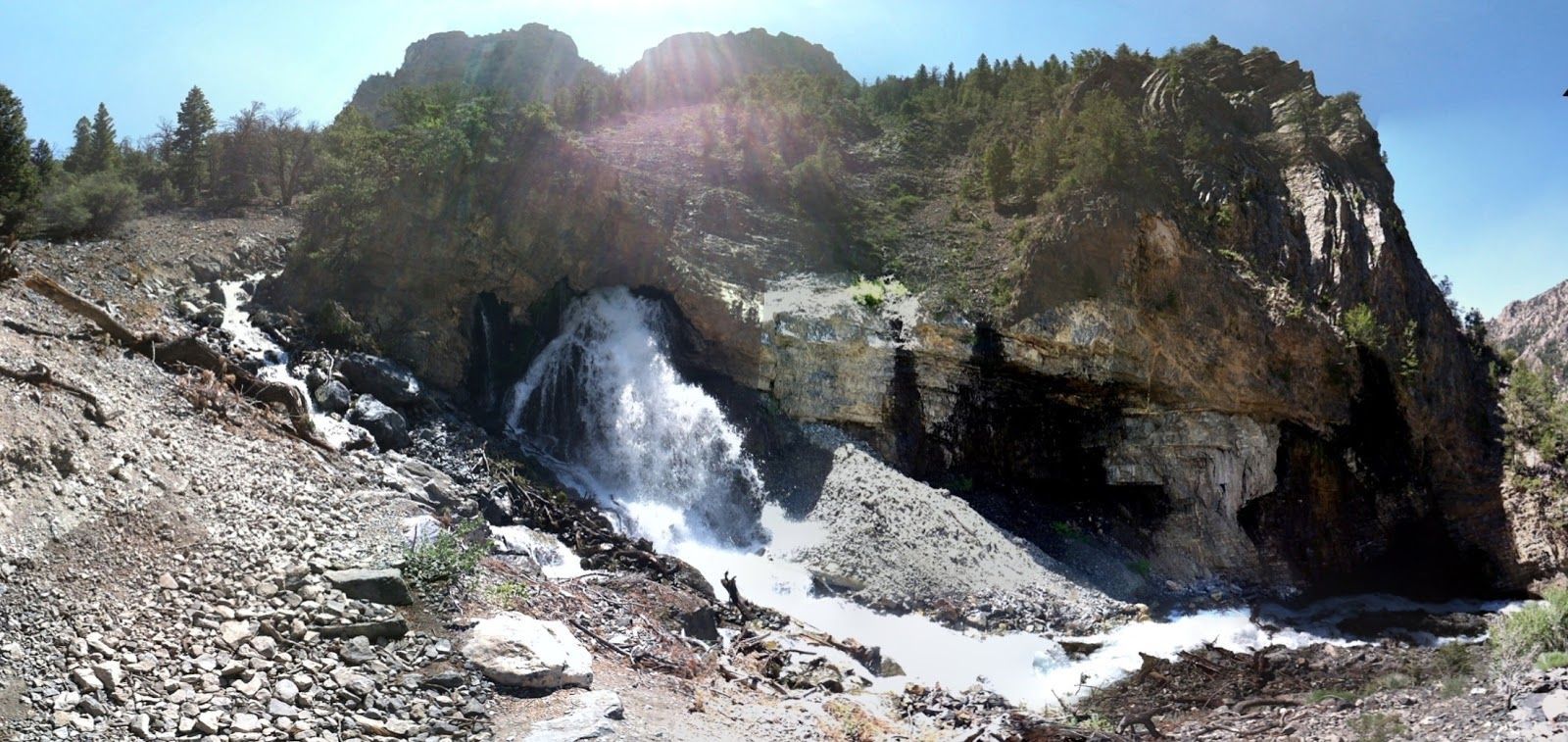 A powerful waterfall gushing from a rocky cavern mouth at the base of a forested mountain slope on a bright, sunny day.