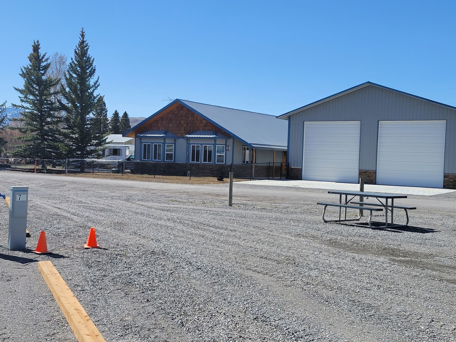 A gravel area with two orange traffic cones, a picnic table, and two buildings under a clear blue sky.