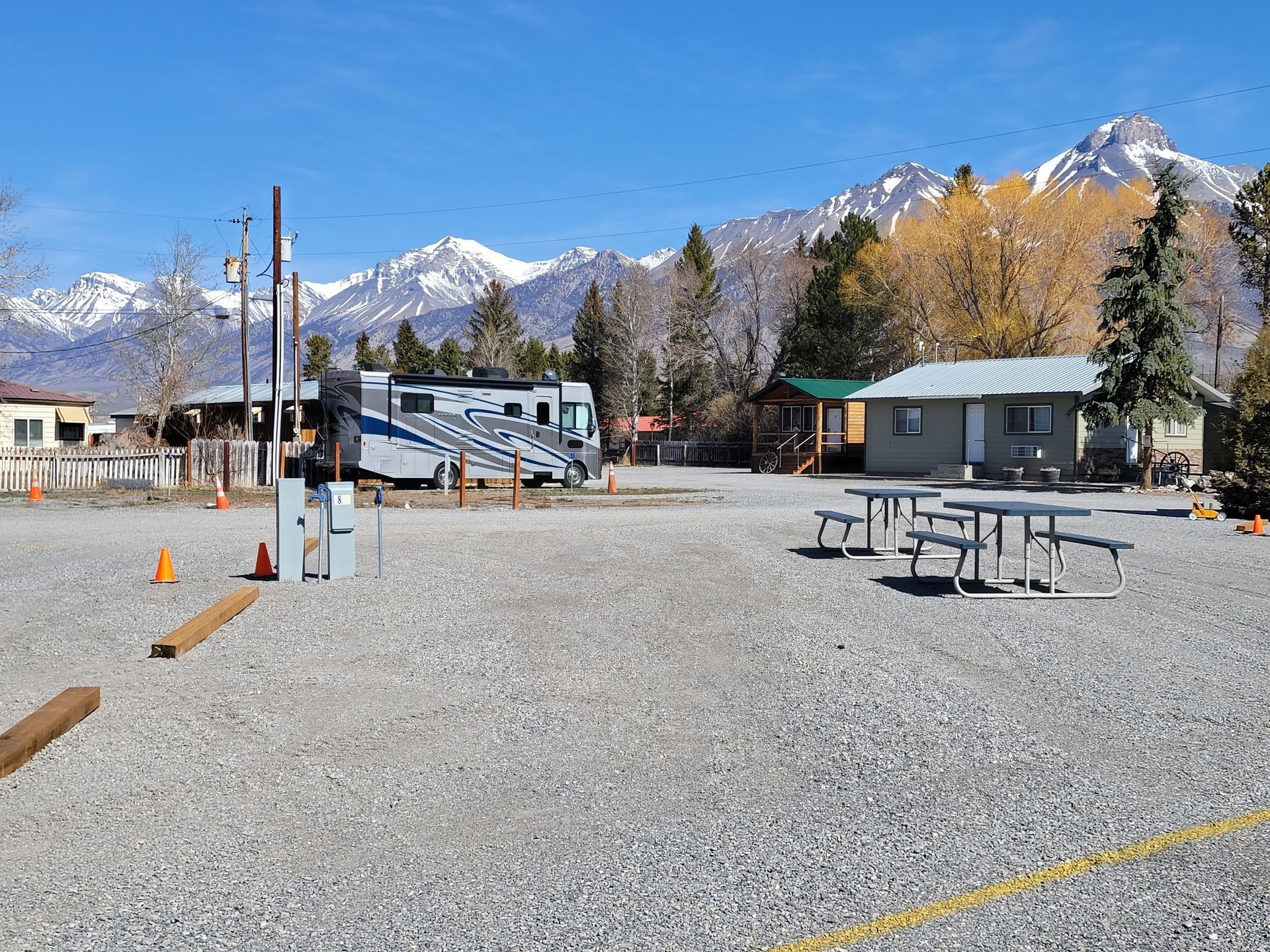 A gravel campground with a parked RV, picnic tables, and a small building against a backdrop of snow-capped mountains.