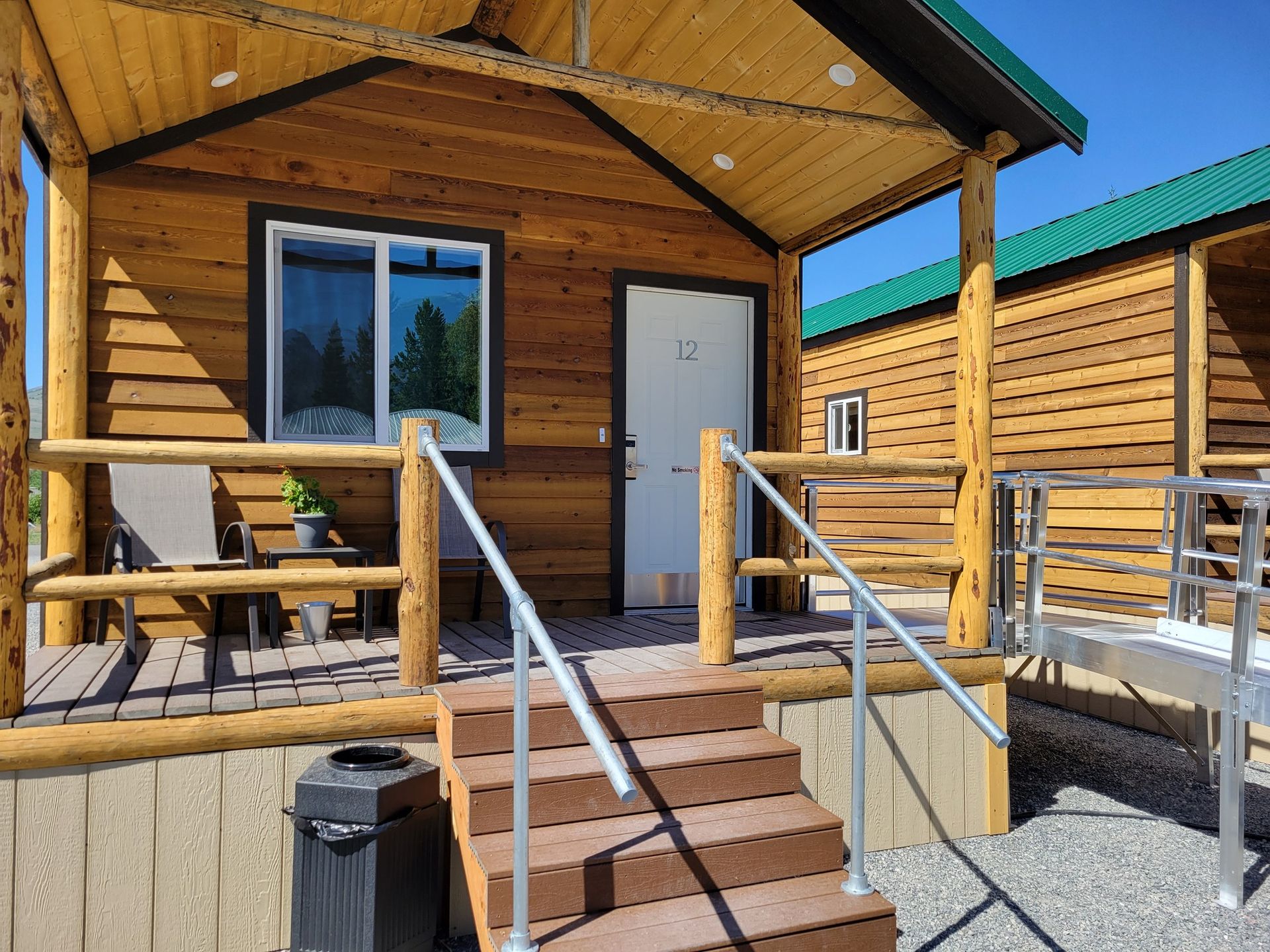 Wooden cabin with a porch, a wheelchair ramp, and stairs, set outdoors under a clear blue sky.