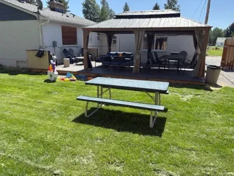 A picnic table sits on a green lawn in front of a house deck featuring a gazebo with patio furniture.