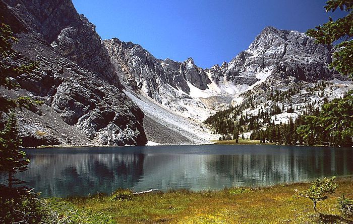 A calm mountain lake reflects rugged, snow-dusted granite peaks under a clear blue sky, framed by trees and grassy banks.