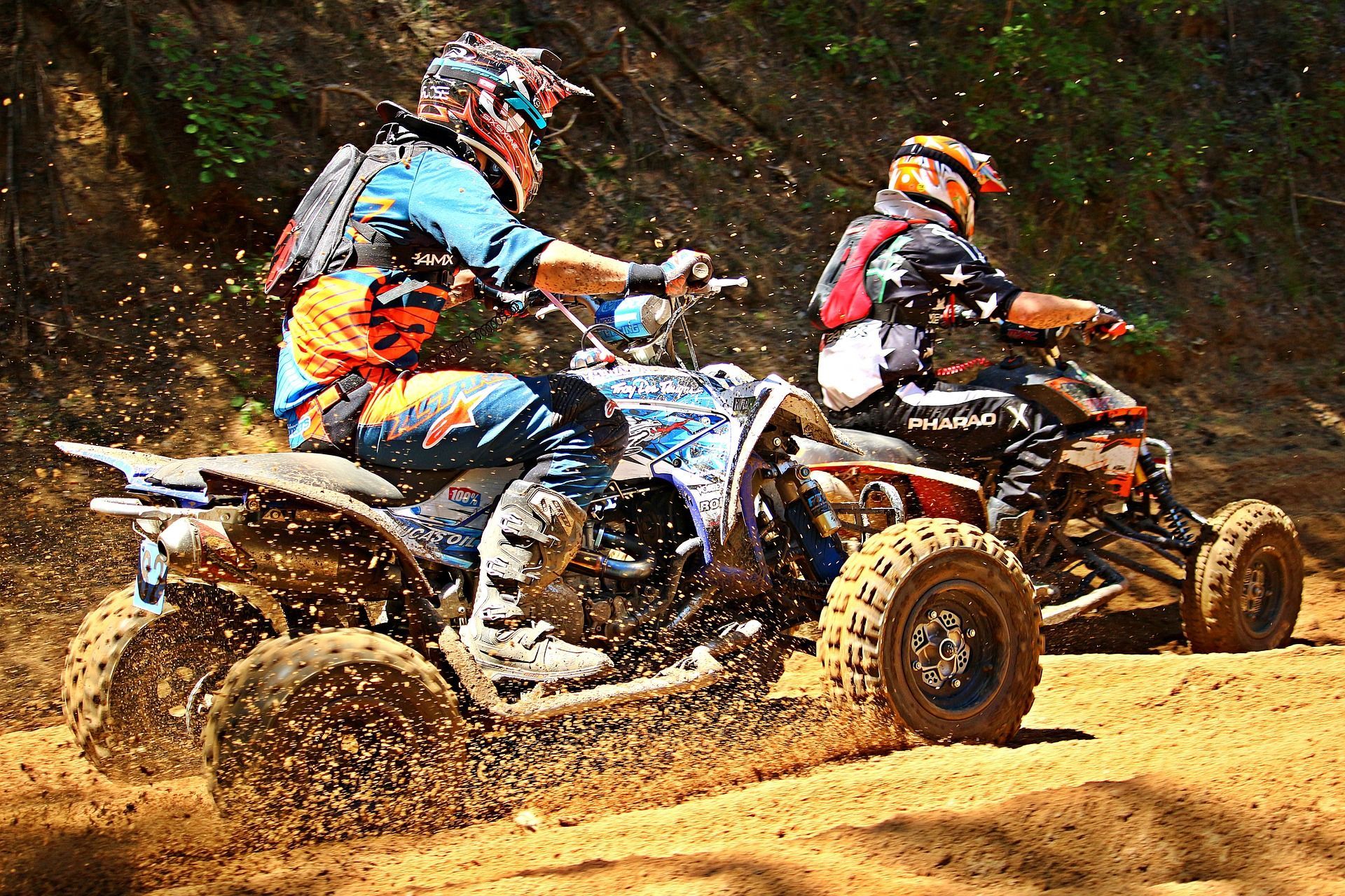 Two people riding ATVs on a dirt track, kicking up mud.