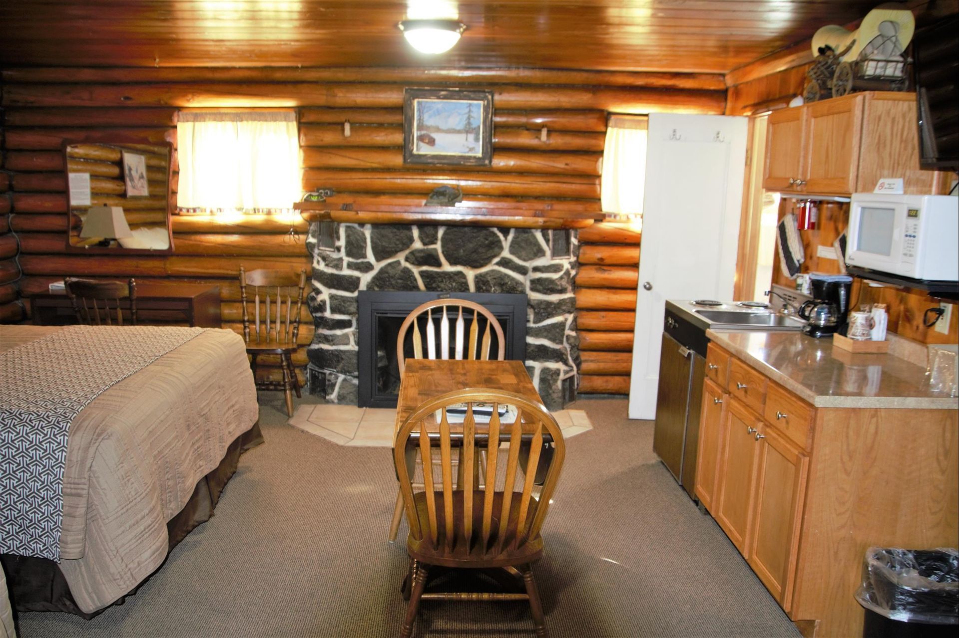 A rustic log cabin hotel room featuring a bed, a stone fireplace, a small dining table, and a kitchenette.