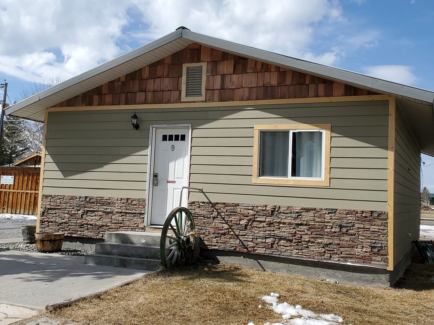 A small, single-story cabin with olive siding, stone veneer accents, wood-shingle gables, and a wagon wheel by the door.
