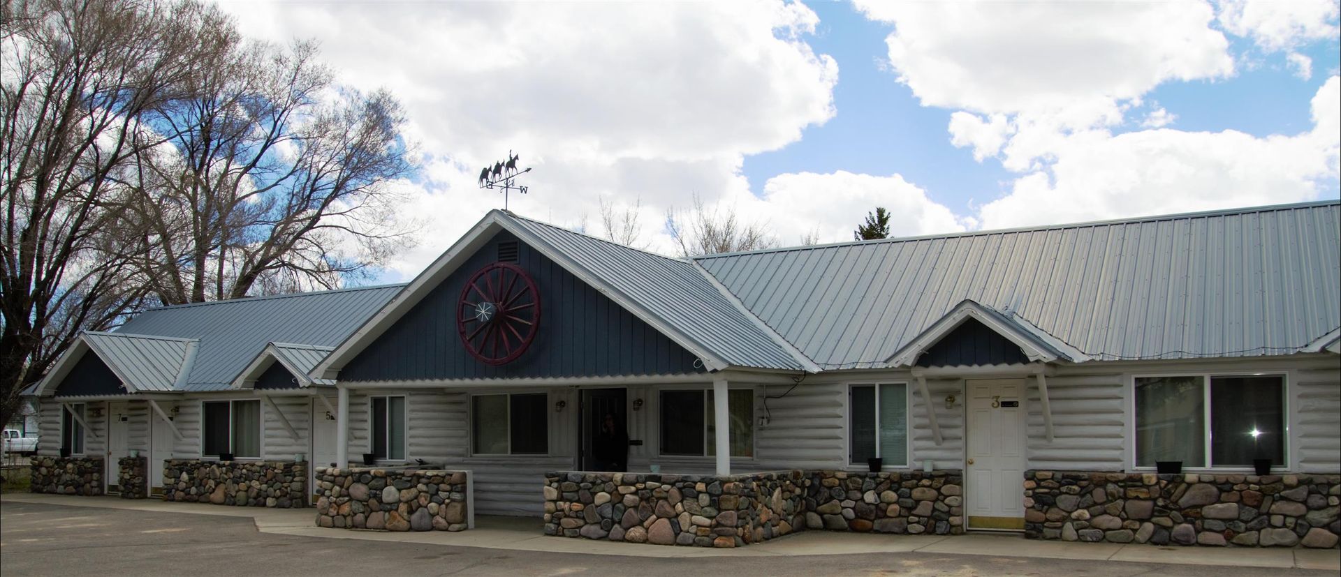A low-slung, white building with a metal roof, stone foundation, and dark blue gables under a blue sky with white clouds.