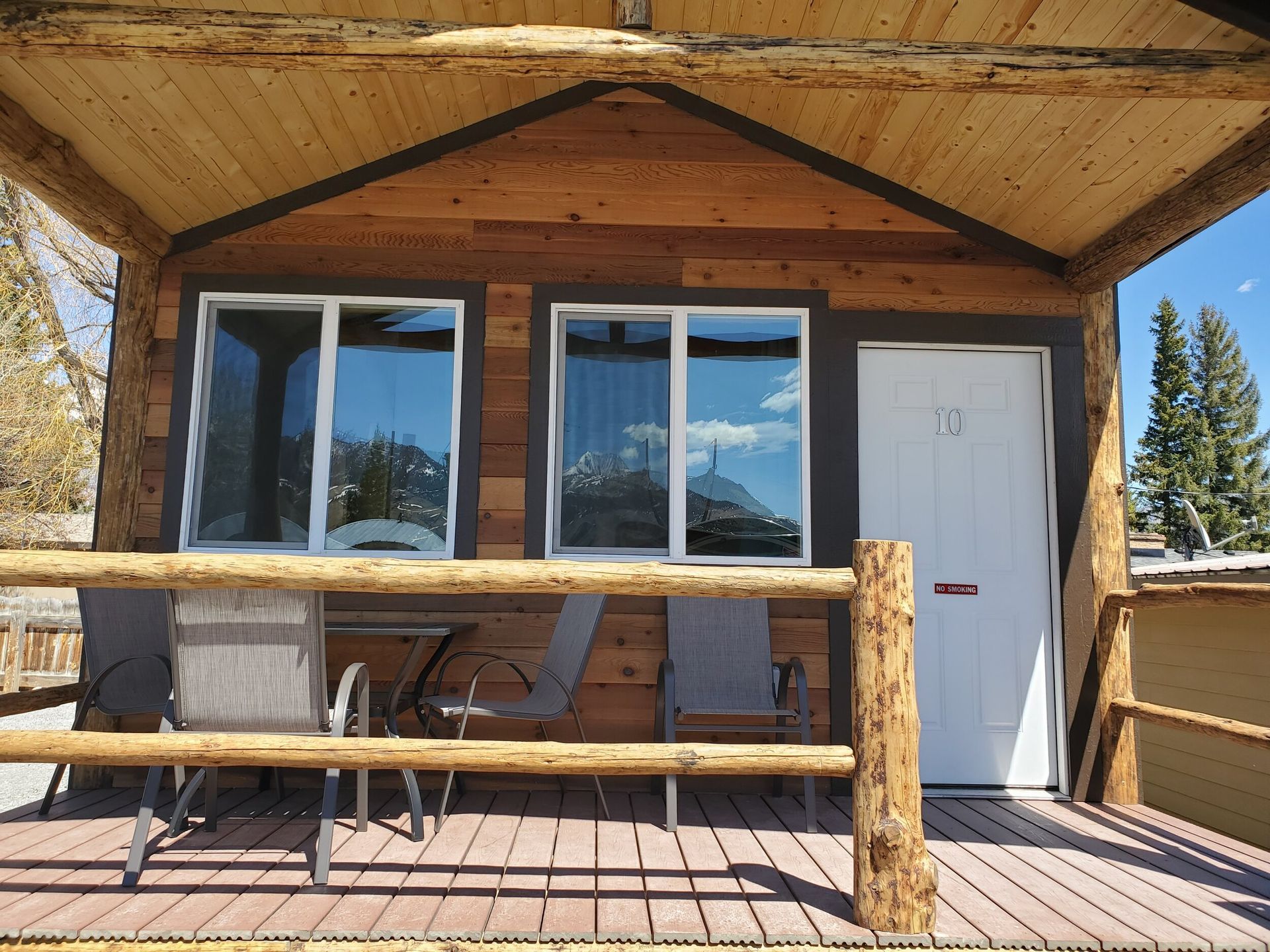 A wooden cabin exterior with a porch, featuring two windows, a white door, and three chairs on a slatted wood deck.