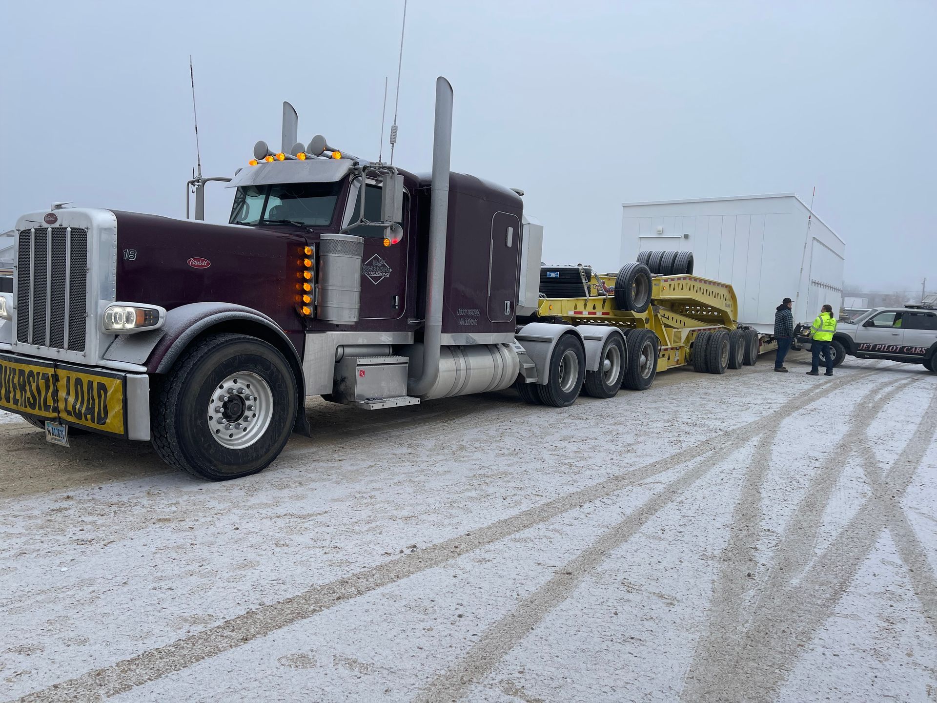 A large maroon truck hauling an oversized load on a snowy road, with workers nearby.