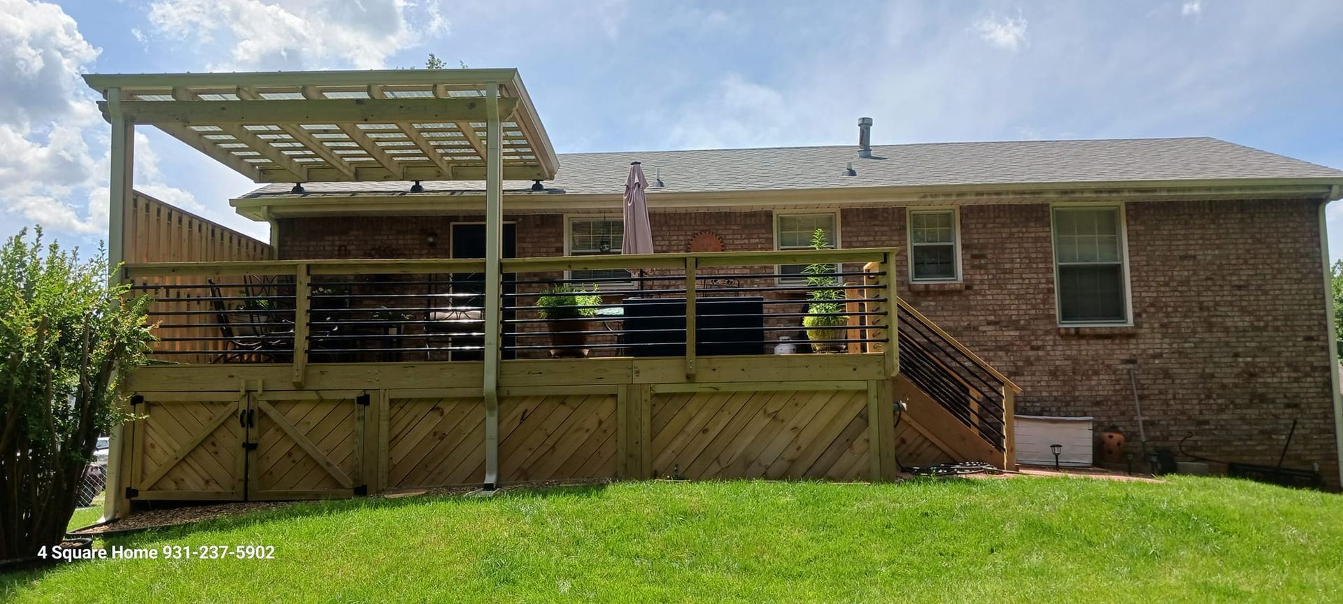 A wooden deck with a pergola is attached to a brick house. Green grass is in the foreground.