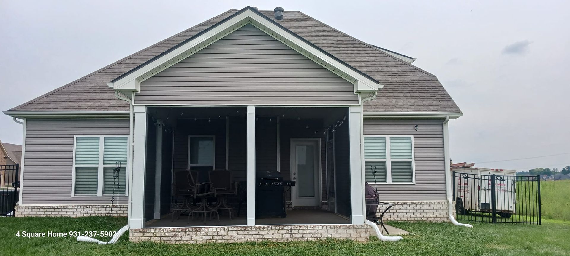 Back of a house with a screened-in porch and grey siding, visible windows, and a field in the background under a cloudy sky.