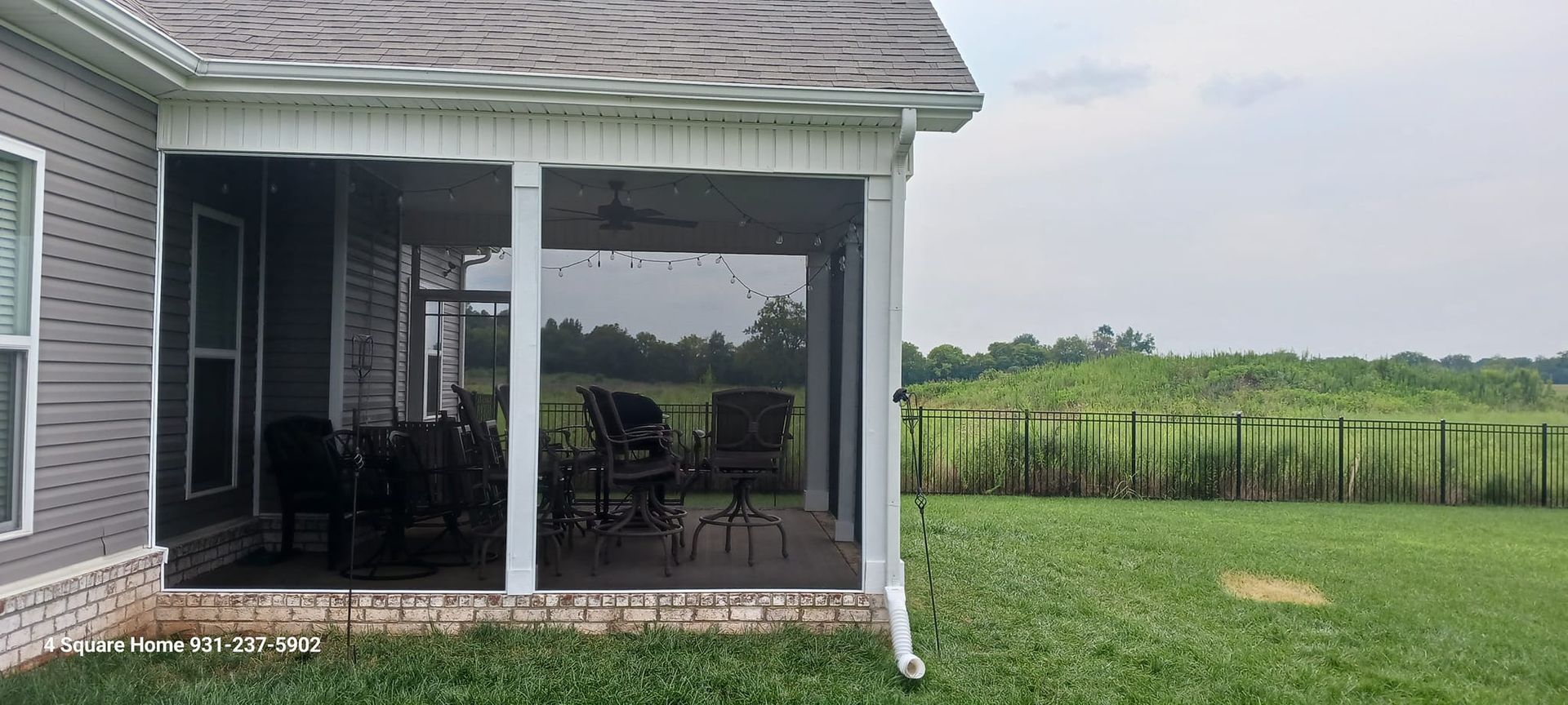 A screened-in porch with outdoor furniture overlooks a grassy field and distant trees under a cloudy sky.