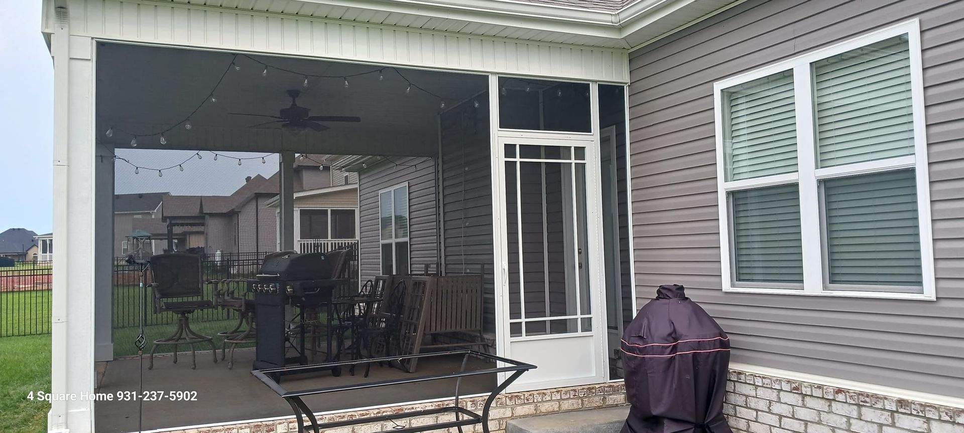 A screened-in porch with outdoor furniture, and a BBQ grill. There's a door, and a gray-sided house with windows. It's raining.