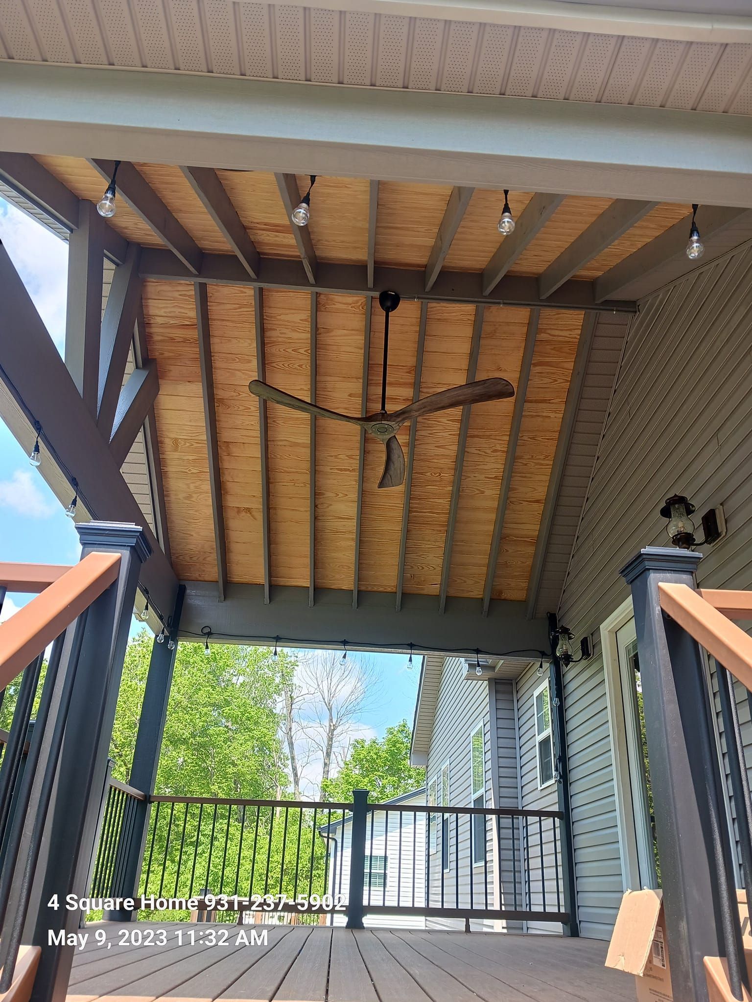 Covered porch with ceiling fan, string lights, and wooden deck. View toward trees and building.