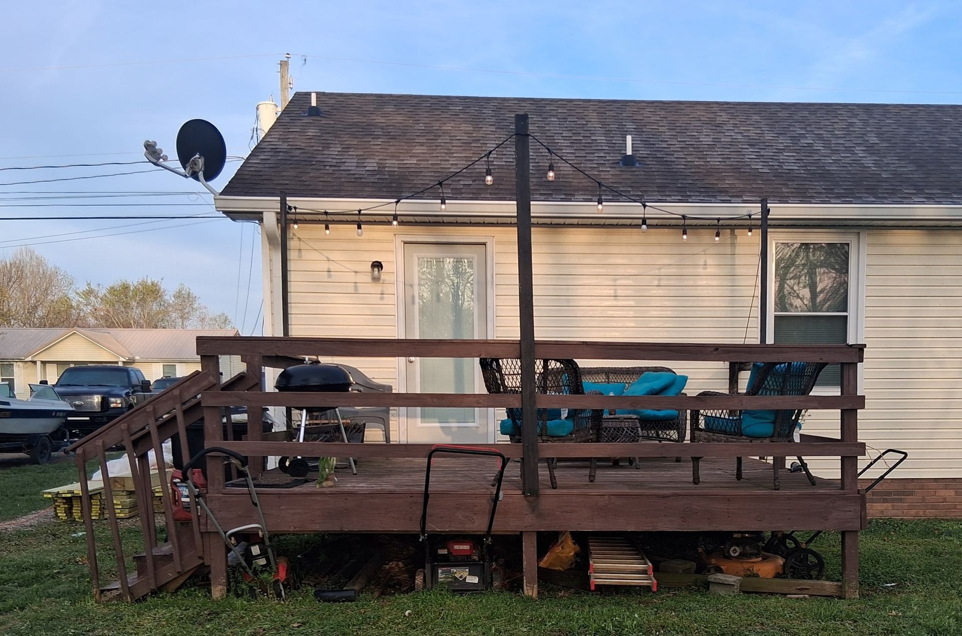 A backyard deck with seating, grill, and string lights against a white house and blue sky.