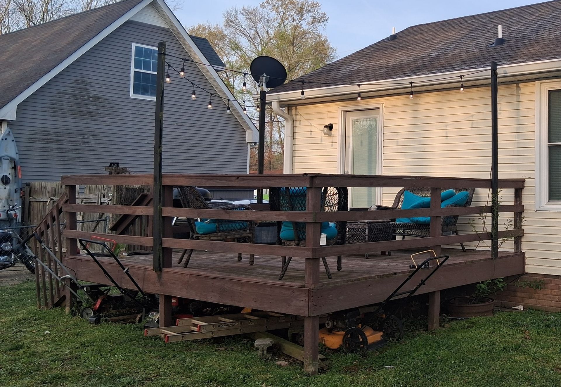 Wooden deck with railing, string lights, and patio furniture, adjacent to a beige house and a gray house.