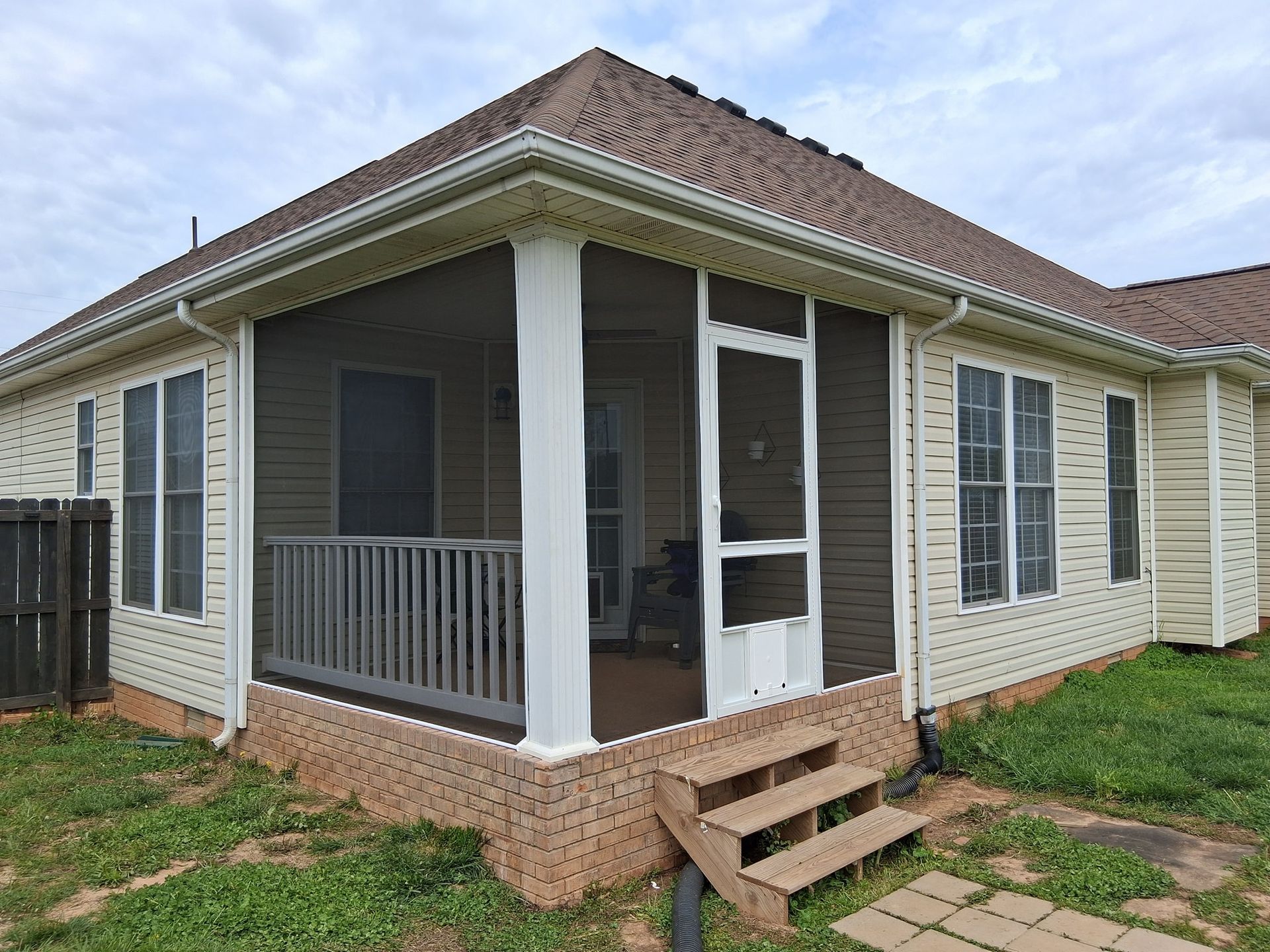 Screened porch with steps, brown roof, and cream siding, next to a house, on a cloudy day.