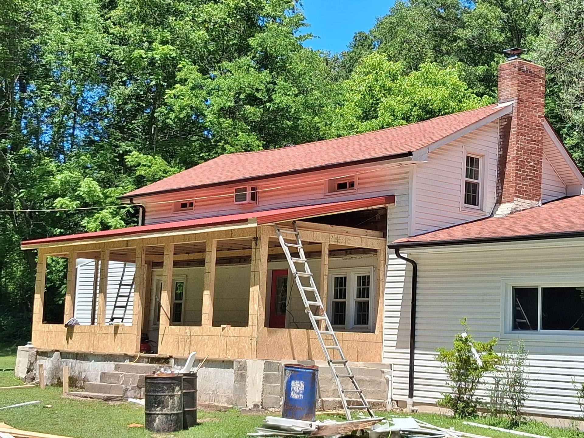 House under construction; porch being added, red roof, ladder, surrounded by trees.