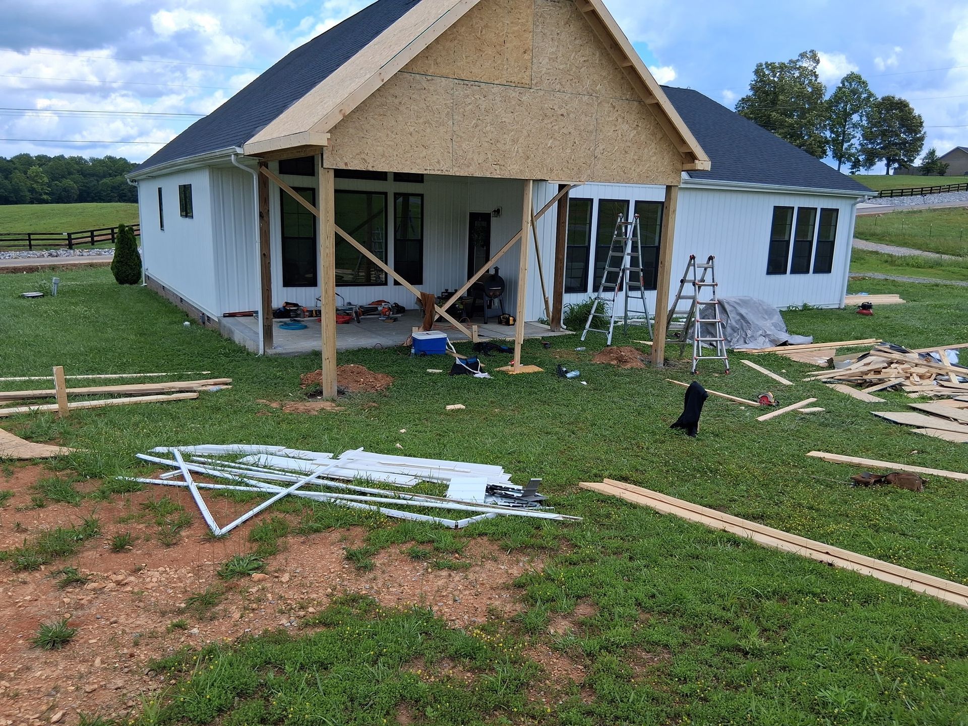 Construction of a covered porch on a white house with a black roof; lumber and tools scattered on the grass.