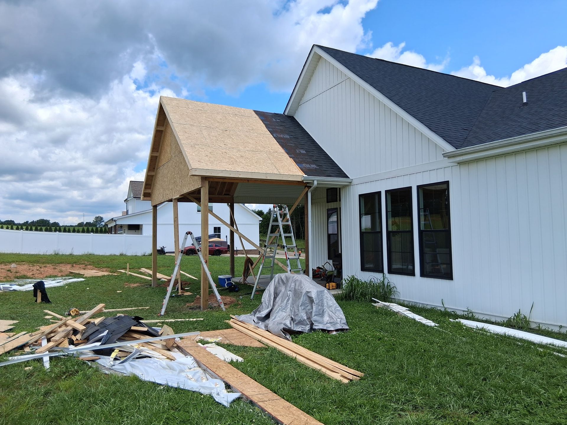 Construction of a porch with a partially completed roof attached to a white house; materials and tools are scattered.