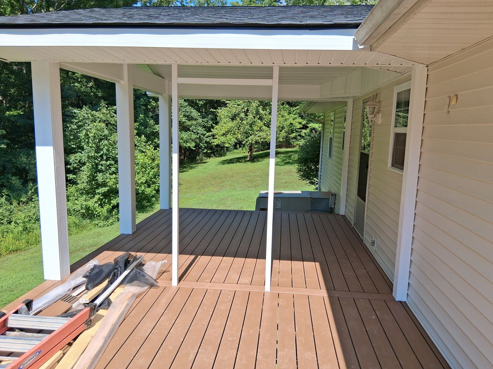 Covered wooden deck attached to a house with white pillars and trim, overlooking a grassy yard.