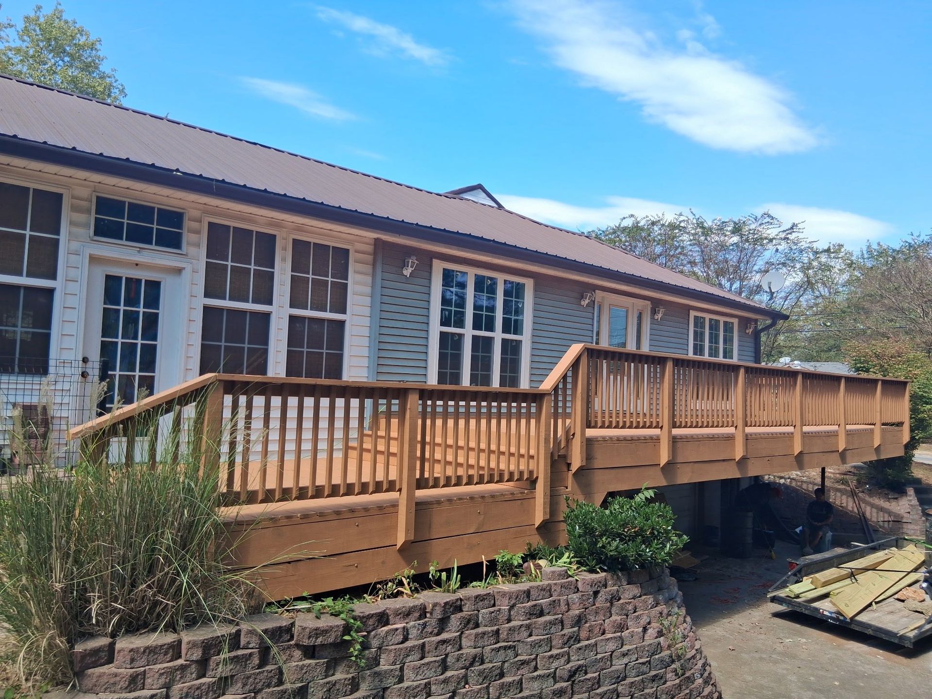 Wooden deck attached to a light blue house with multiple windows and a brown roof. Sunny day.