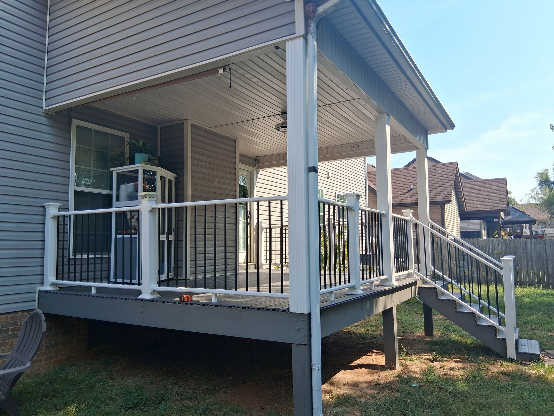 Covered deck with gray and white railings, steps, attached to a house with a backyard.