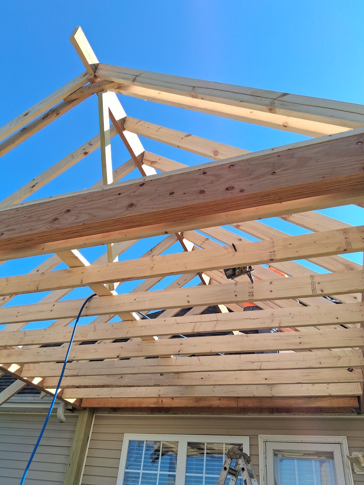 Wooden roof structure under construction against a blue sky, above a house with windows.