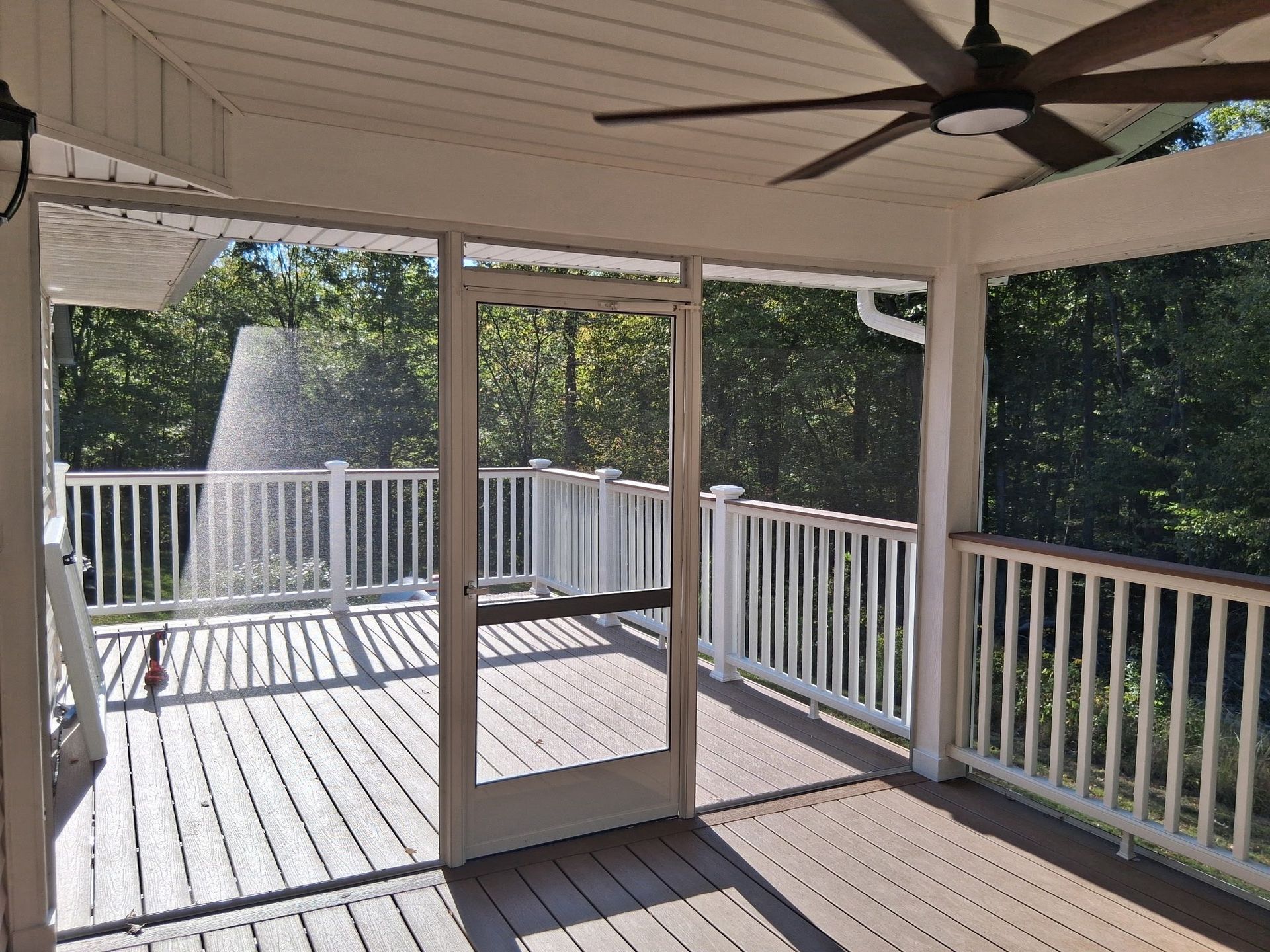 Screened porch with wooden deck, white railing, and ceiling fan; a misty sprinkler in the background.