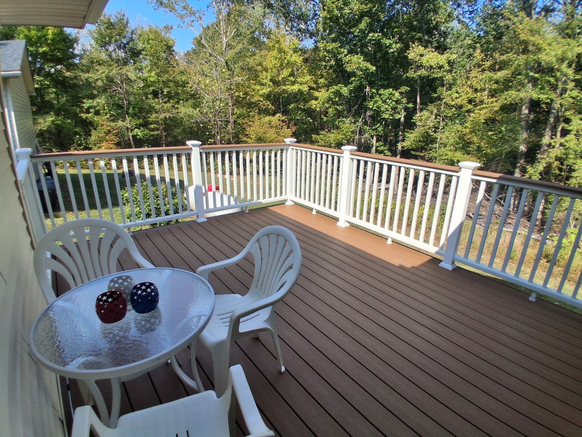 Brown deck with white railing, small table with chairs. Trees in the background.