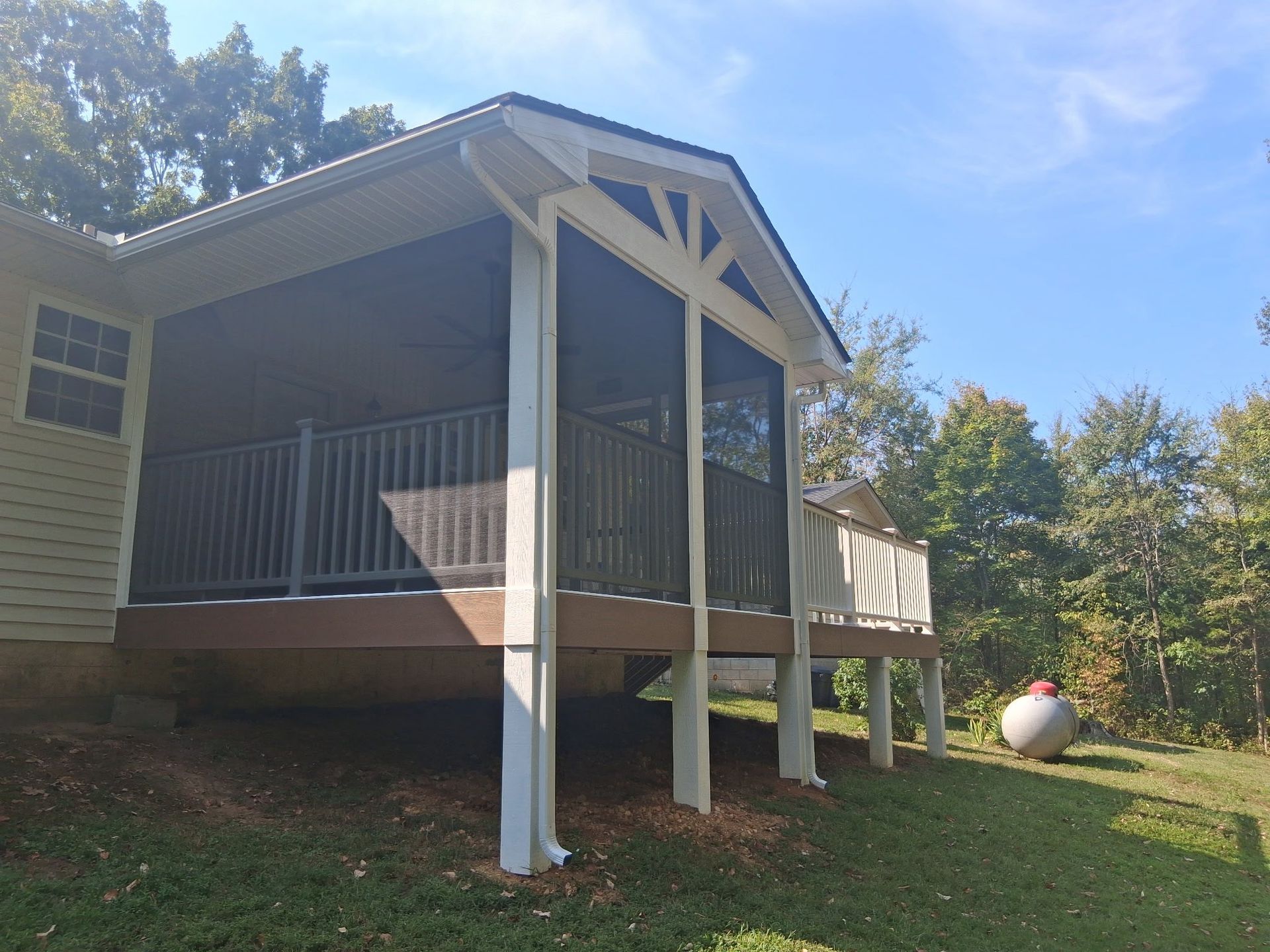 Screened porch attached to a house with white columns, brown deck, and propane tank on the grass.