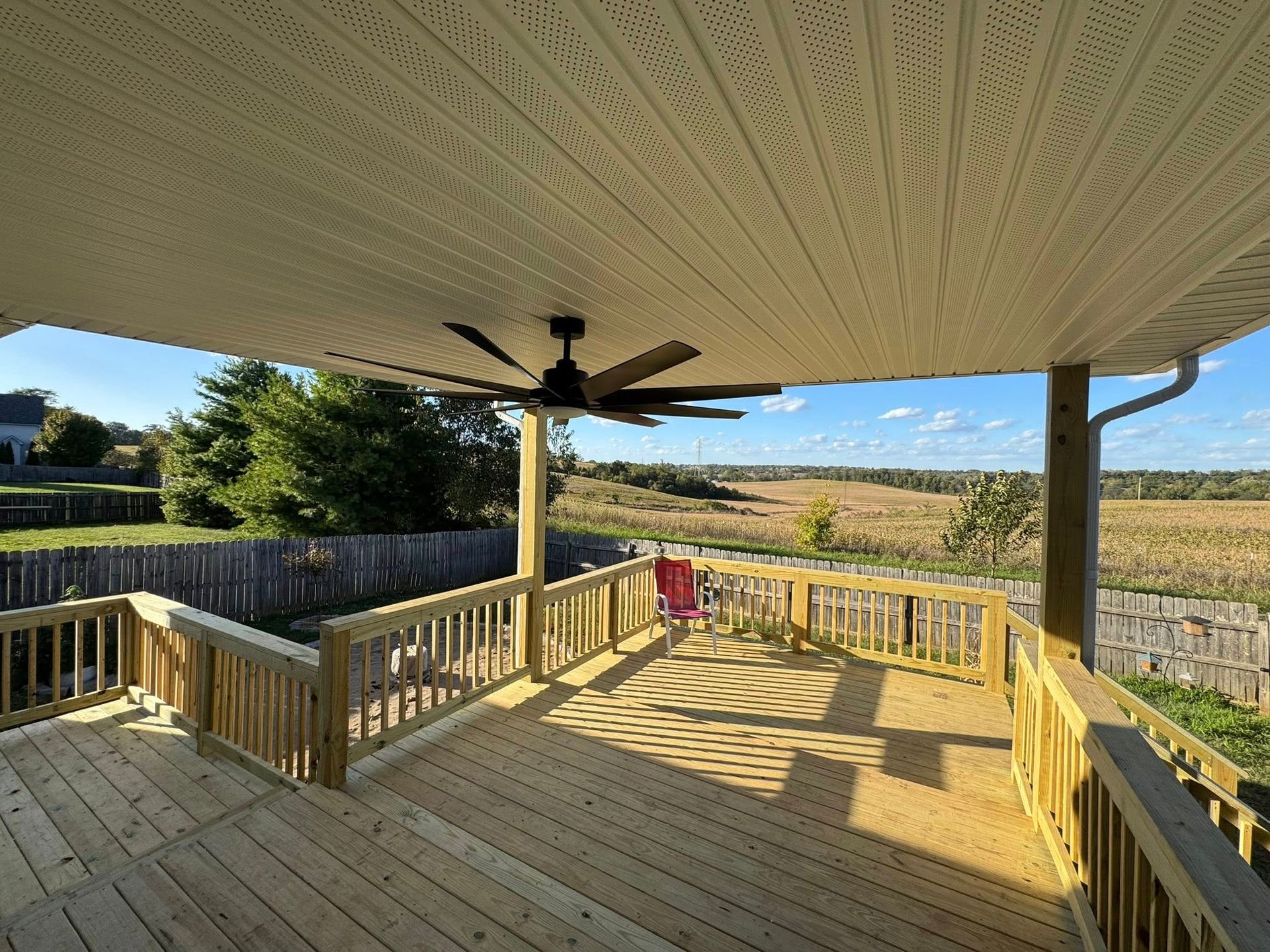 Wooden deck with railing and ceiling fan, overlooking a field.