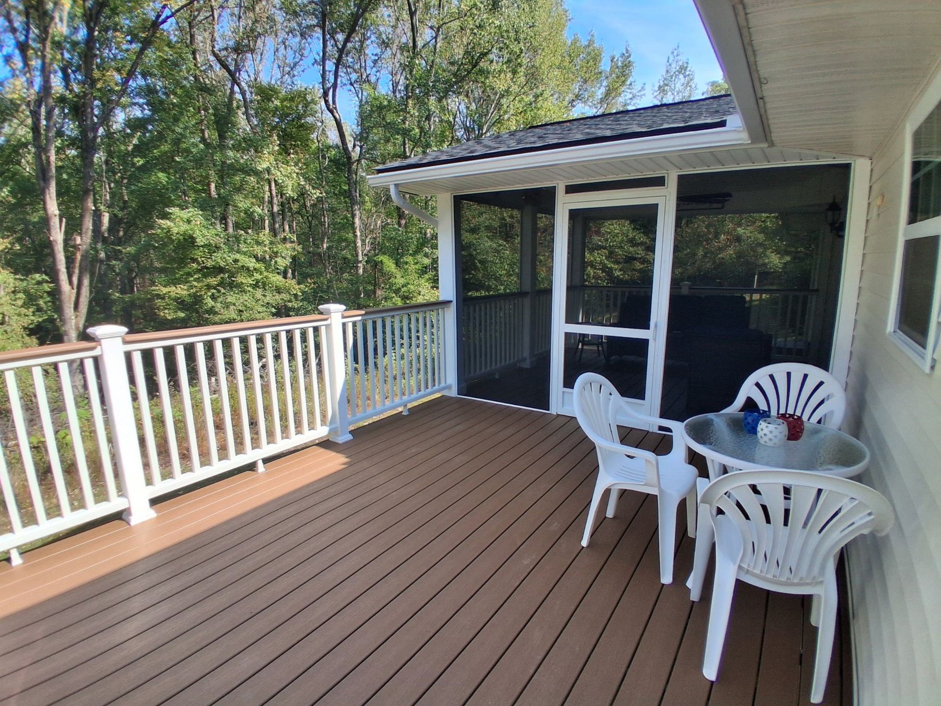 Deck with white railing, screen porch, brown composite decking, and plastic chairs overlooking trees.