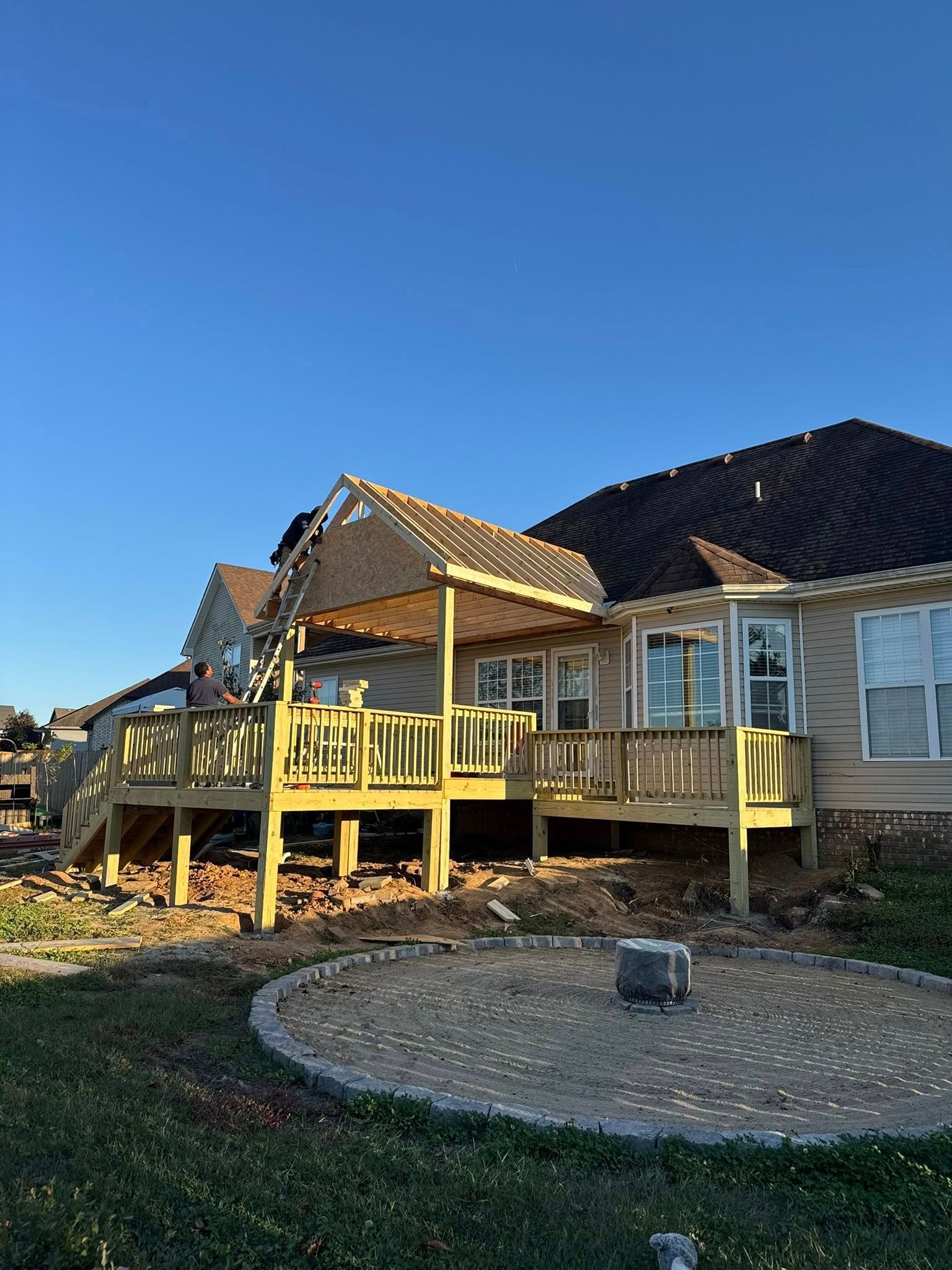 Wooden deck with a roof extension under construction, next to a brick-sided house, clear blue sky.