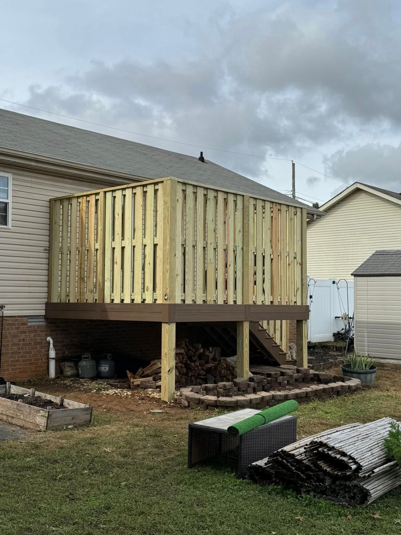 Wooden deck with vertical fencing, attached to a house. Exterior with cloudy sky and yard debris.