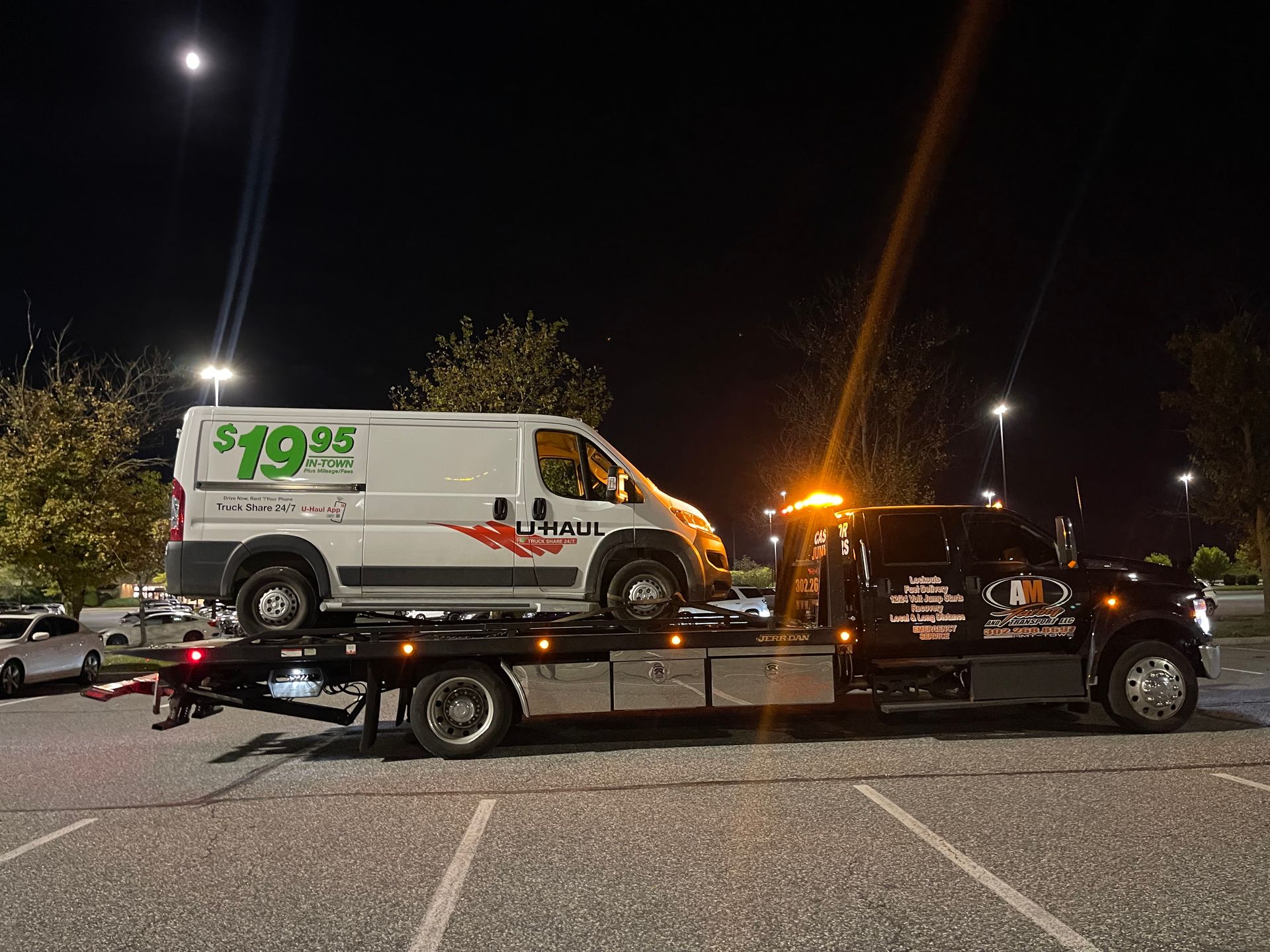 Tow truck carrying a white U-Haul van on a dark night, parking lot setting, lit by streetlights.