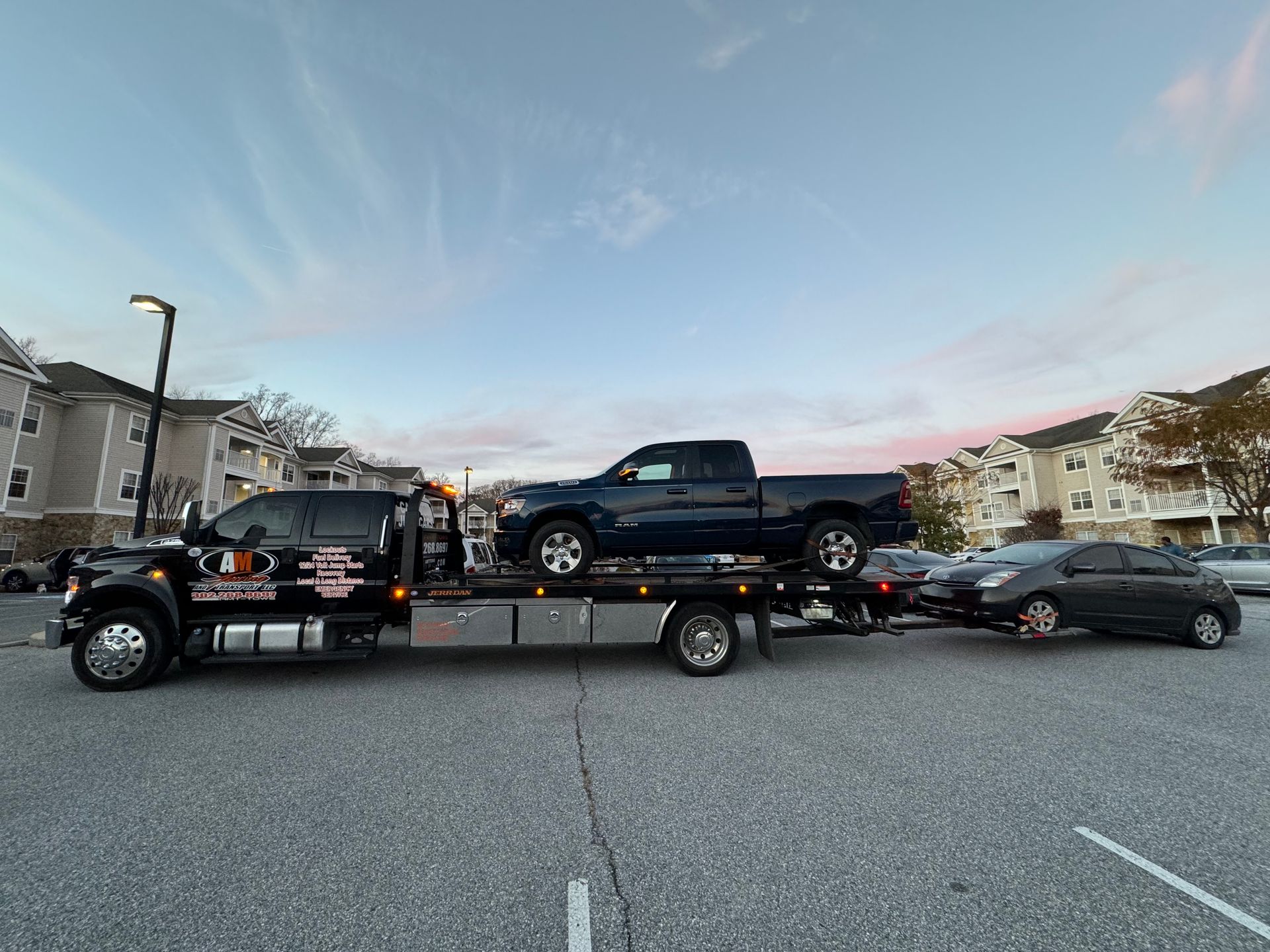 Tow truck with a black pickup truck and a dark gray car on its flatbed, in a parking lot.