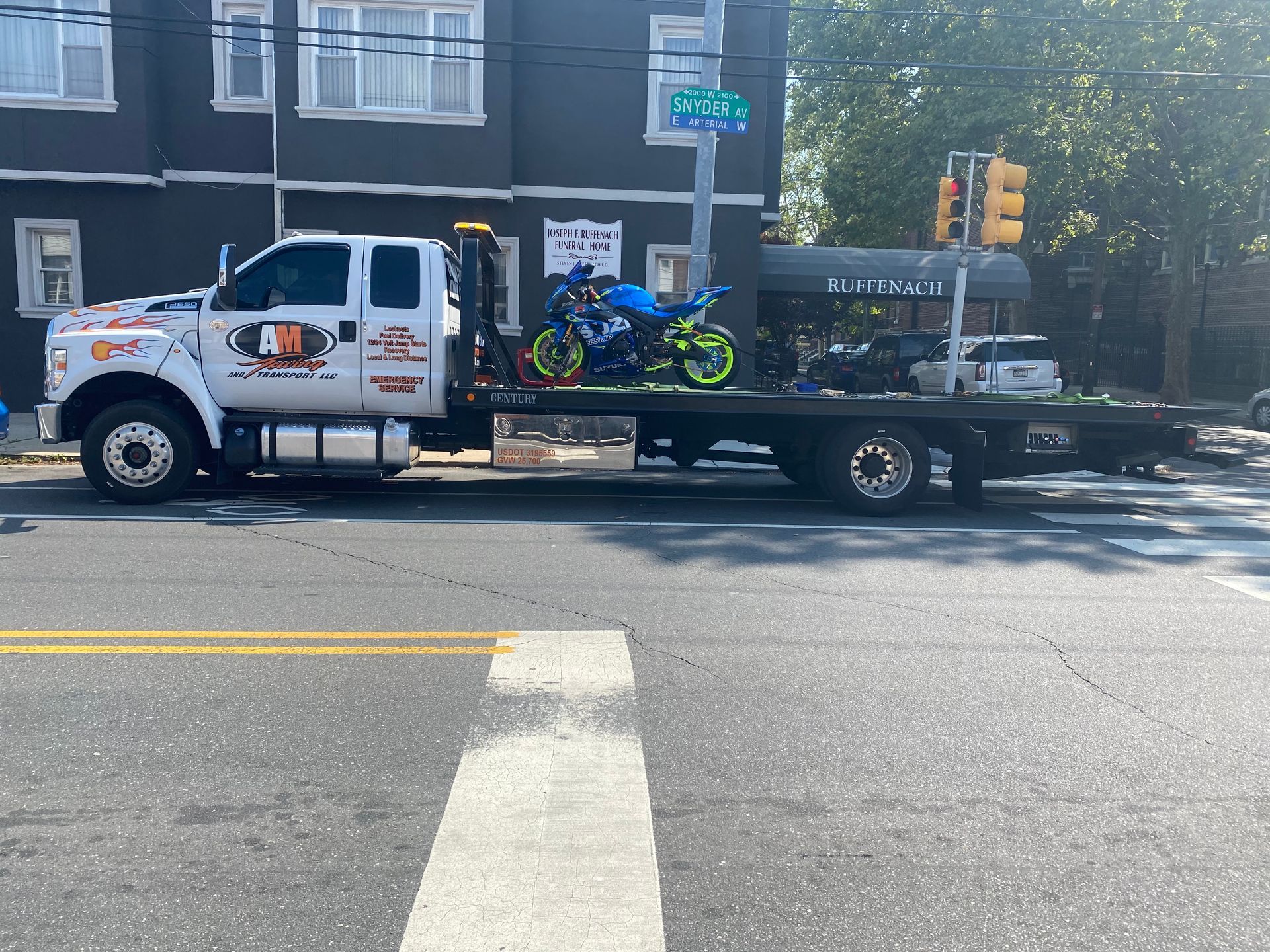 Tow truck carrying a motorcycle on a city street. Building and traffic light visible.