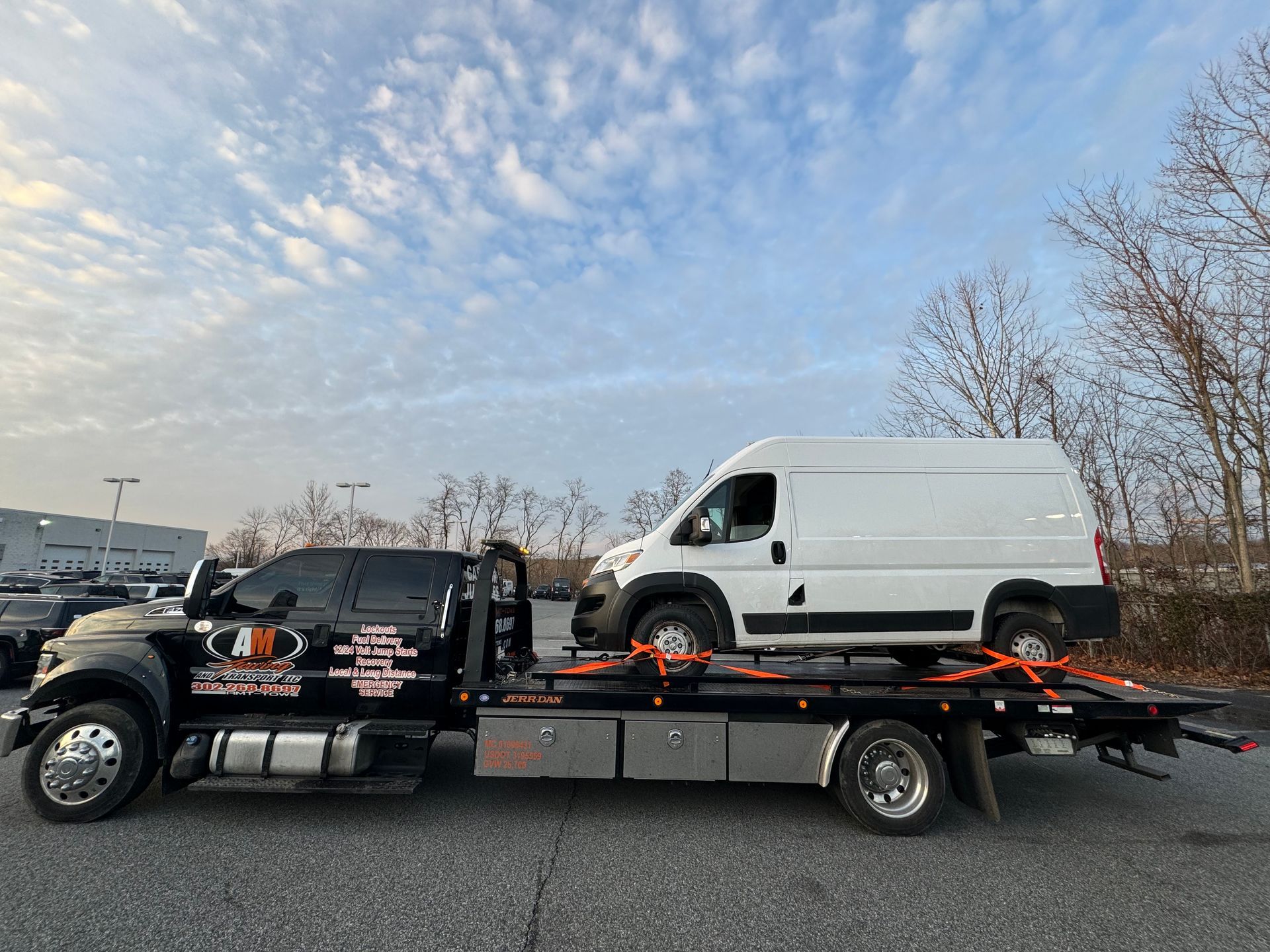 Tow truck carrying a white van on its flatbed. Outdoors on a cloudy day.