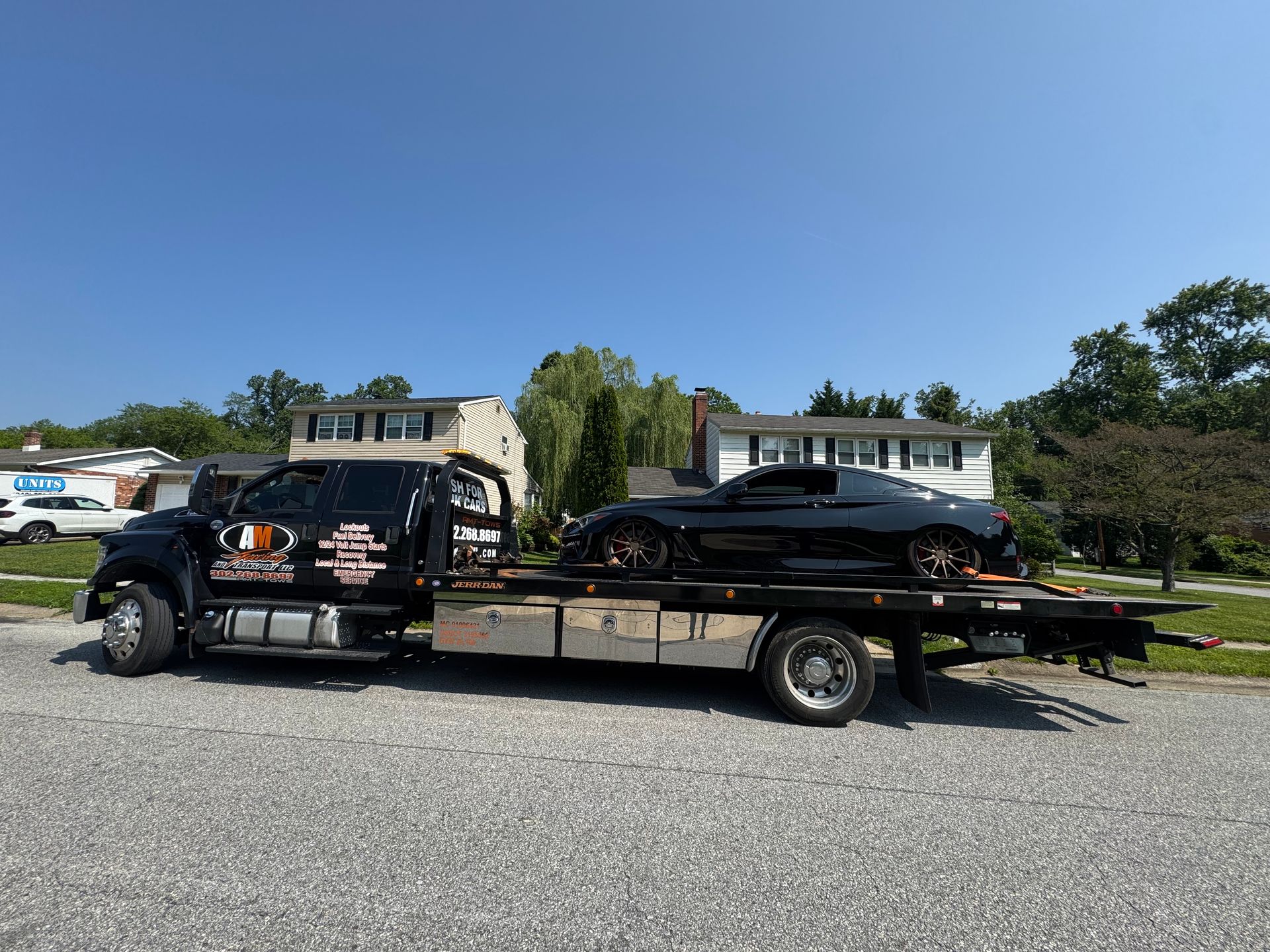 Black tow truck carrying a black sports car on a residential street under a blue sky.
