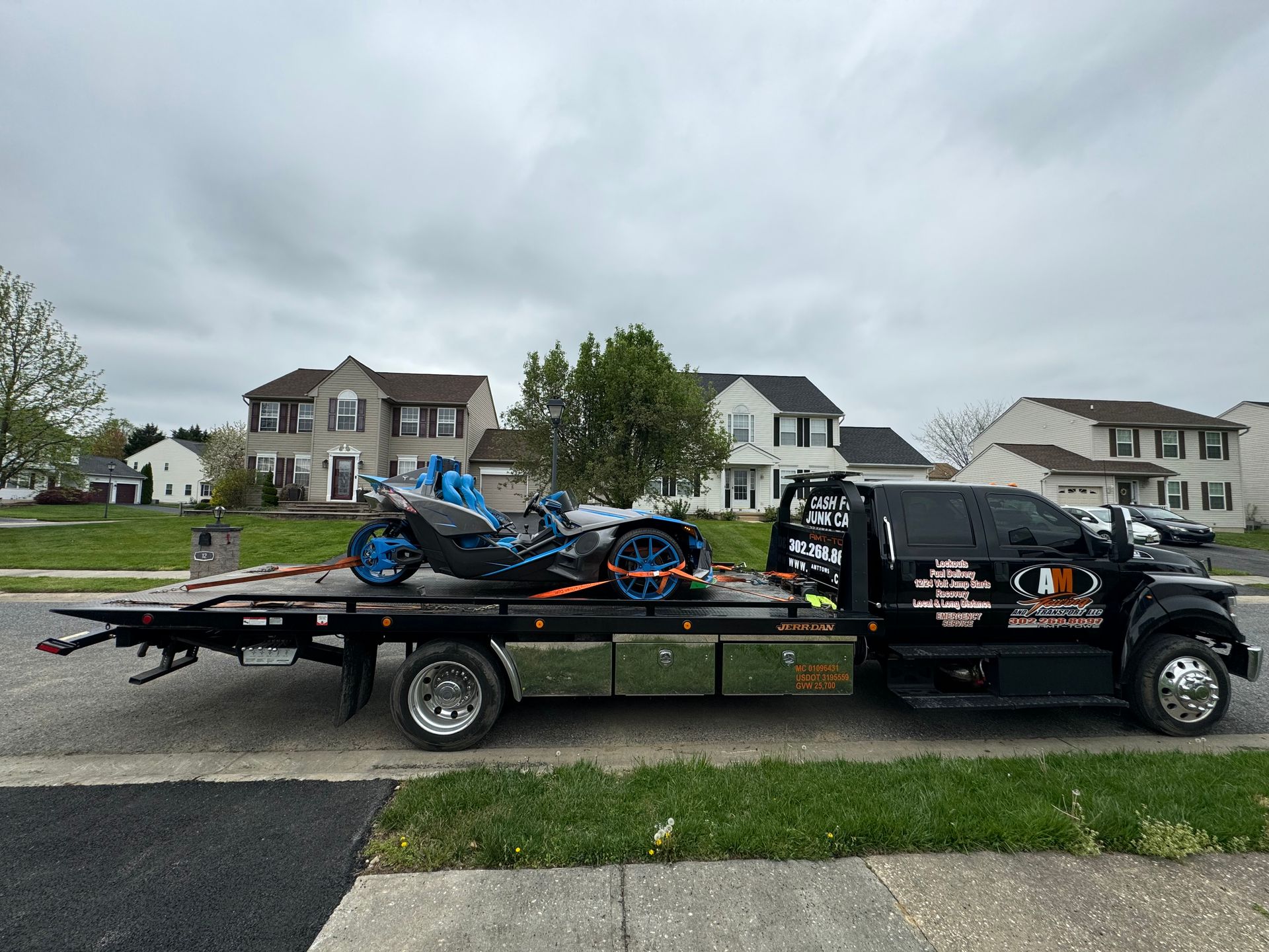 A black tow truck with a blue and black Slingshot on its flatbed, in front of suburban houses.