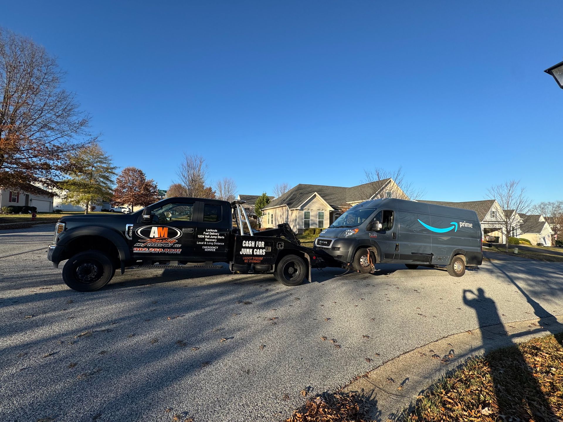 Tow truck towing an Amazon delivery van on a suburban street under a blue sky.