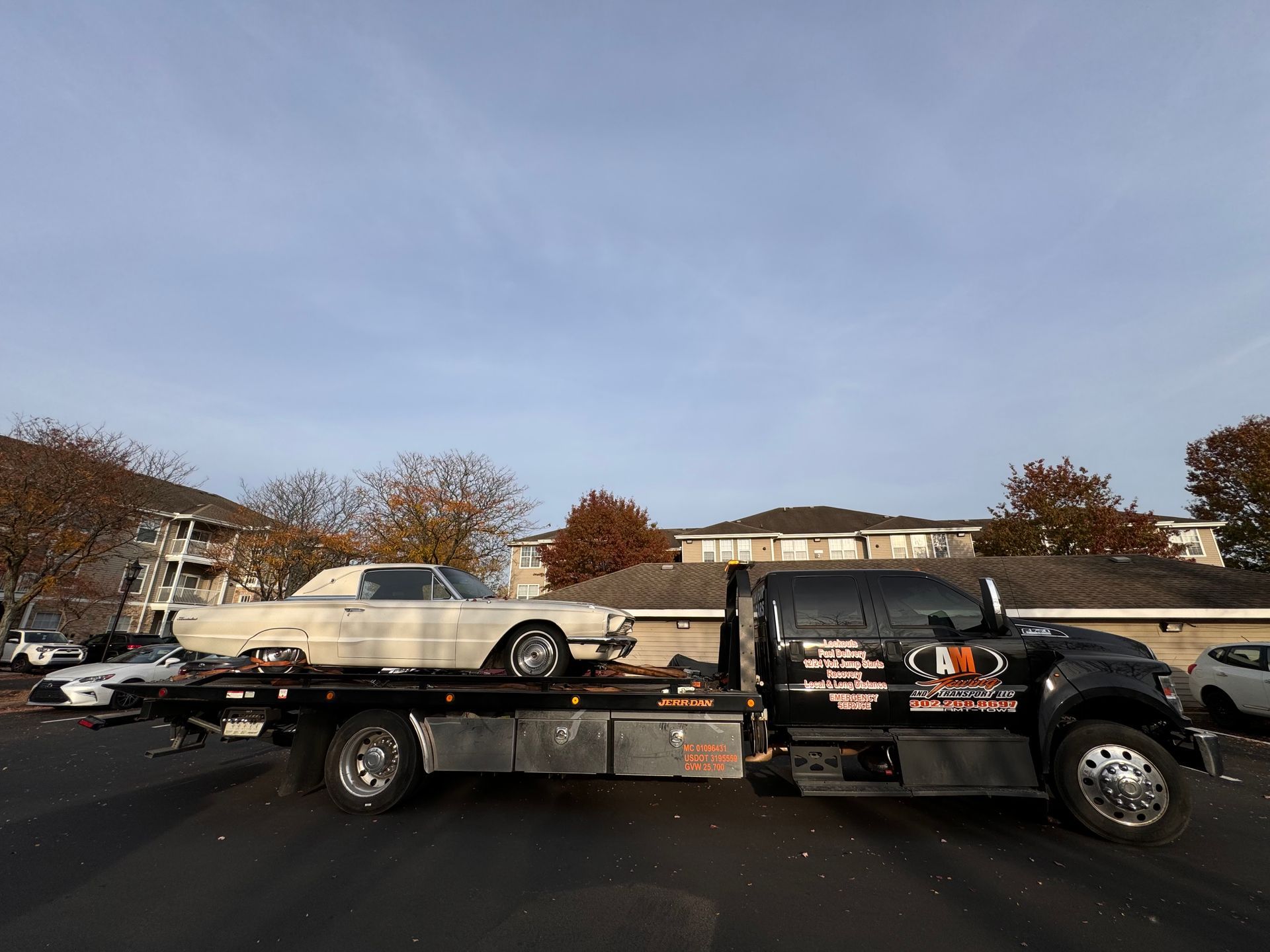 Tow truck carrying a vintage white car. Black truck on asphalt, blue sky.