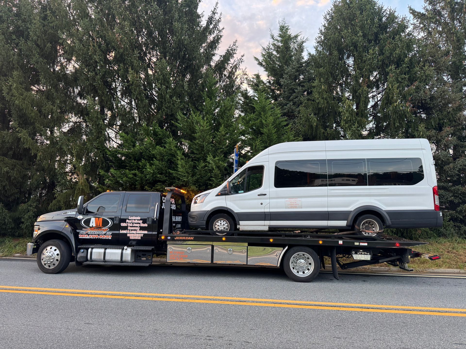 A black tow truck transporting a white passenger van on a roadside with trees in the background.