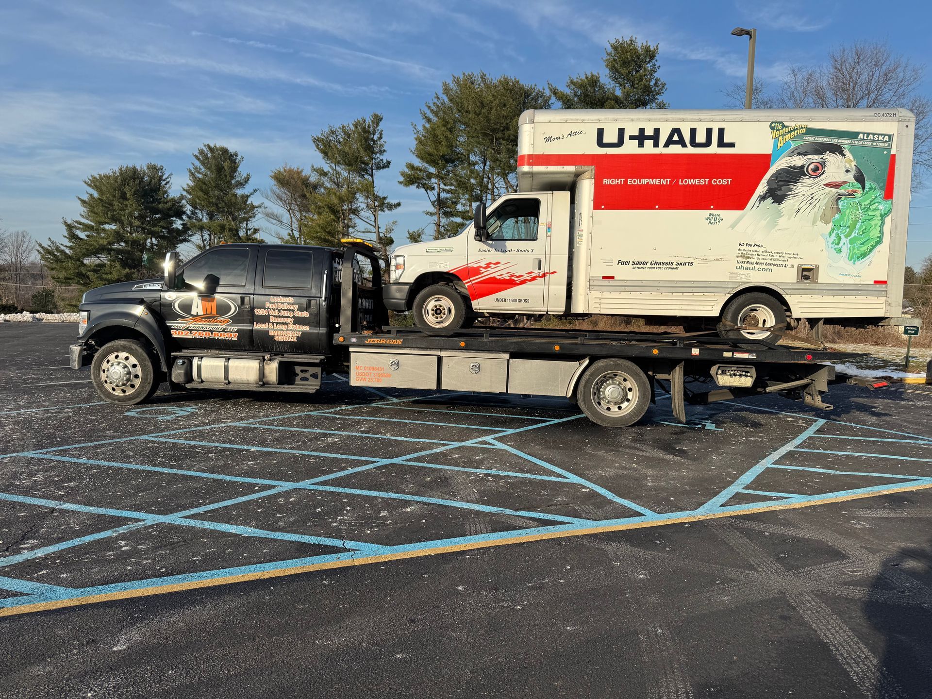 Tow truck transporting a U-Haul truck on a flatbed in a parking lot.