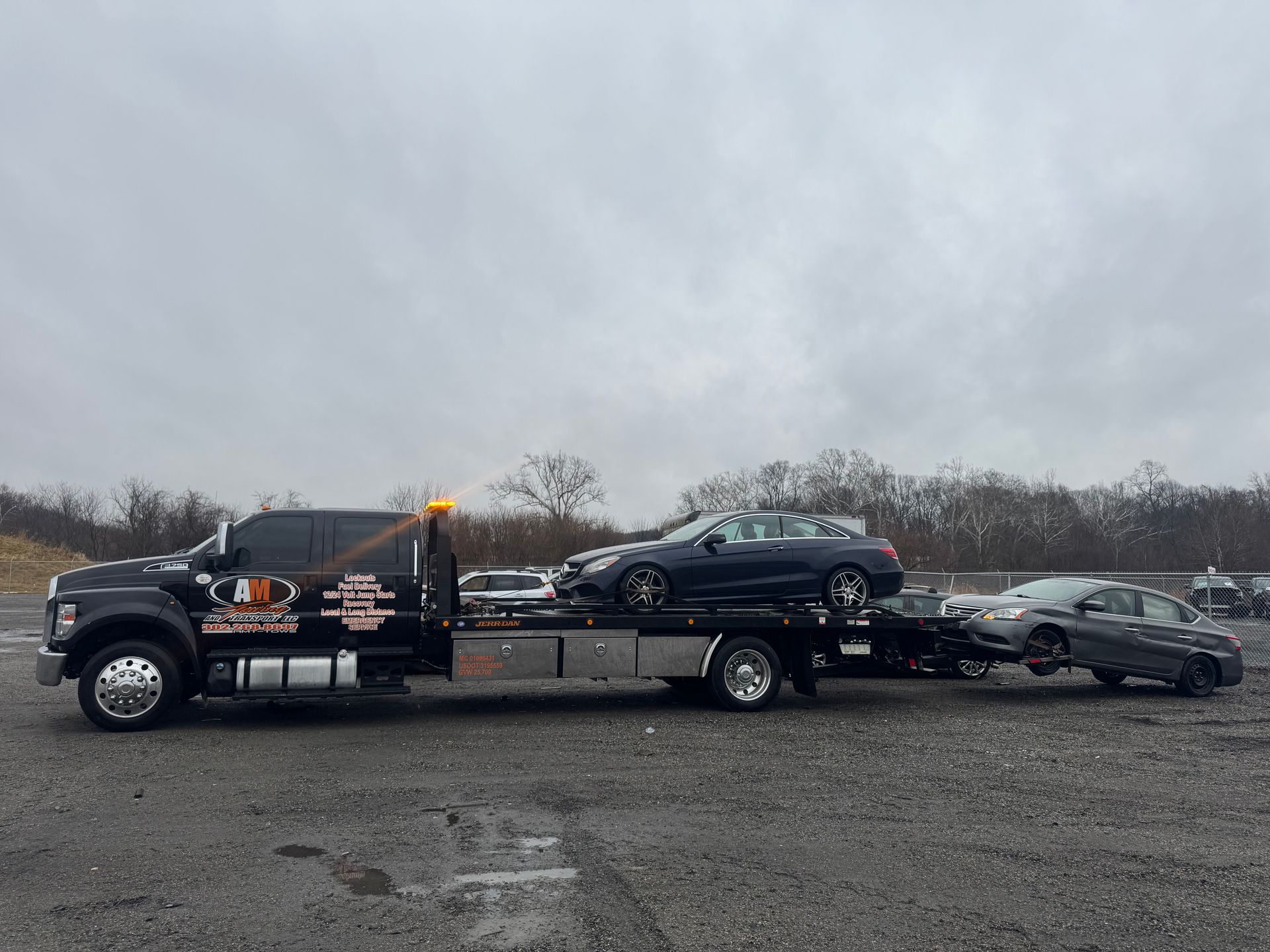 Tow truck carrying two cars on a cloudy day. The truck is black with orange lights and the cars are dark-colored.