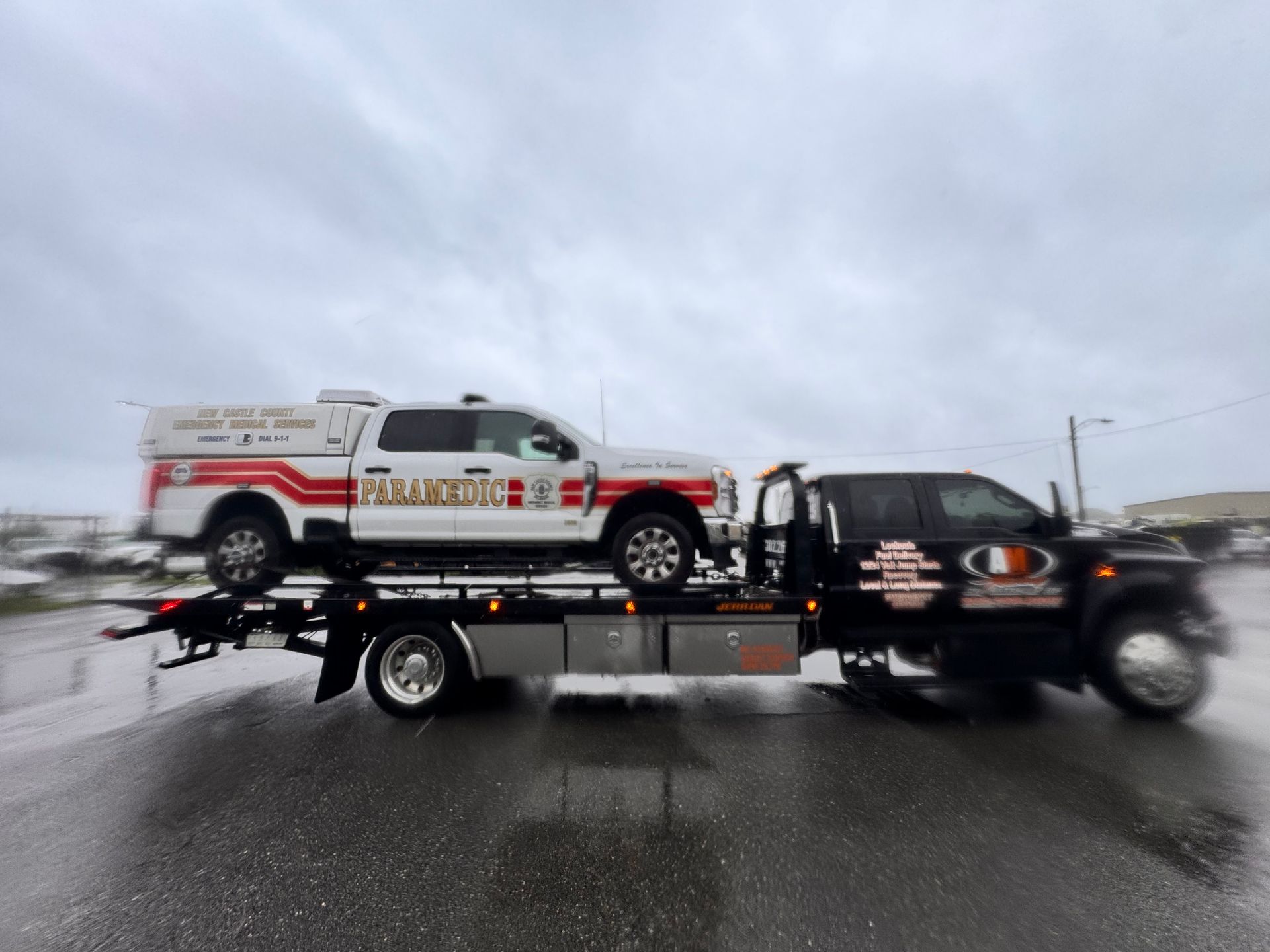 Tow truck carrying a white and red service truck under a cloudy sky.
