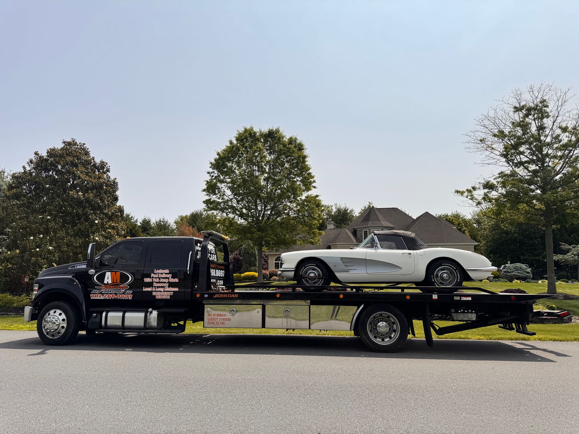Tow truck with a white vintage Corvette on its flatbed. Sunny outdoor setting.